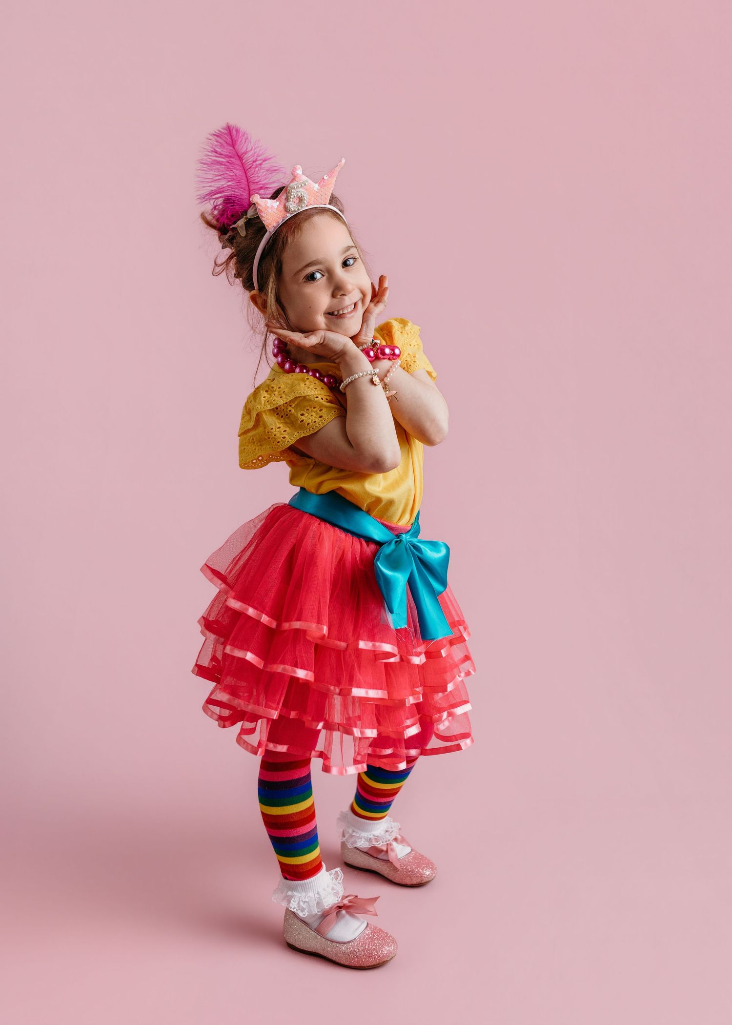 Child wearing colorful tutu dress and striped leggings twirls against pink backdrop.