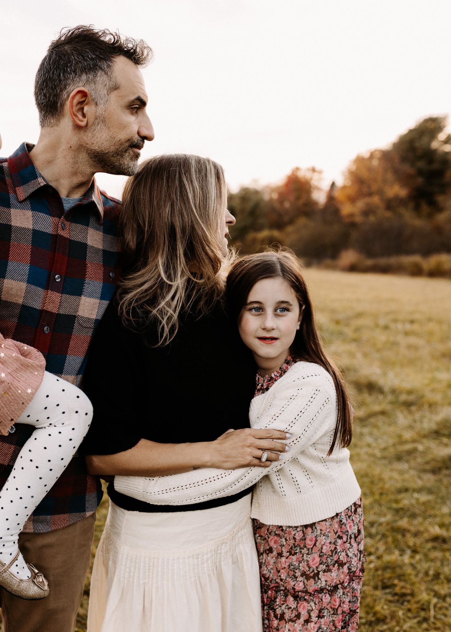 Family of four posing outdoors in a fall field, father holding toddler, mother and daughter embracing warmly.
