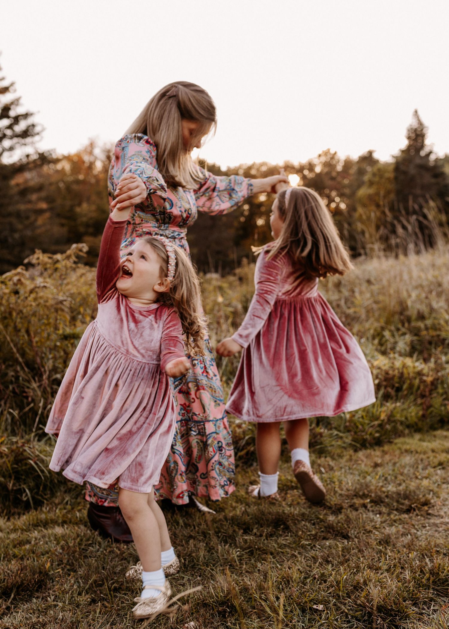 Three young girls in pink dresses playing together in a field during golden hour.