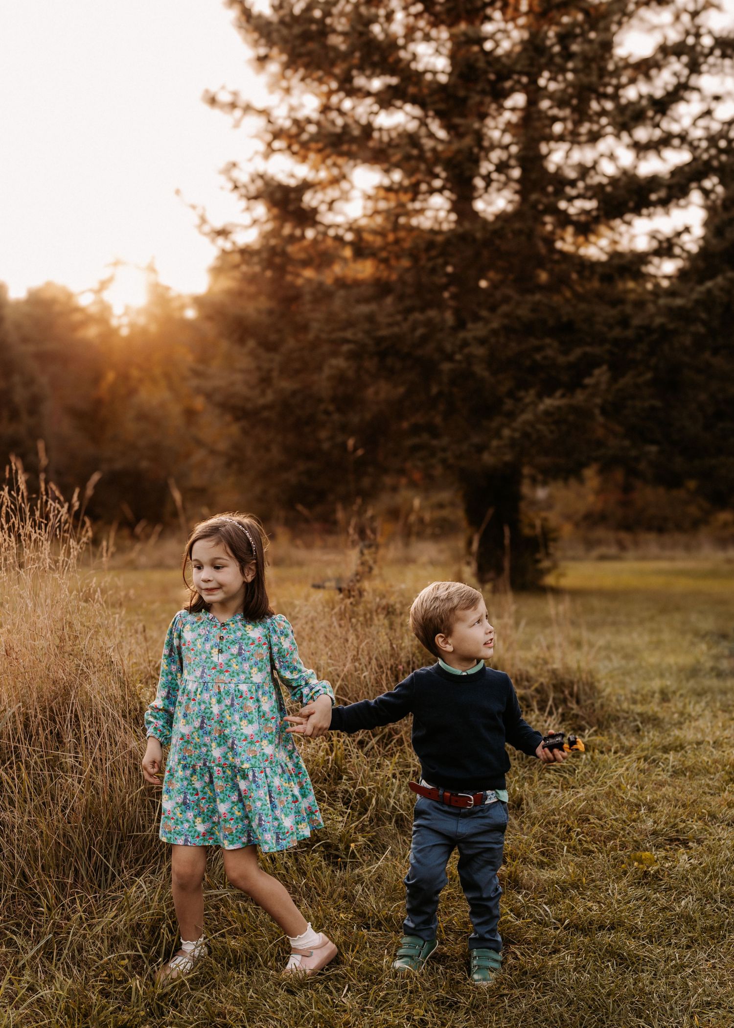 Two young children hold hands in a golden sunset field, girl in floral dress and boy in navy sweater and jeans.