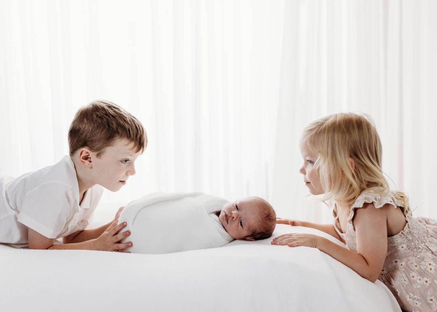 Two children looking at newborn baby lying on white bedding.