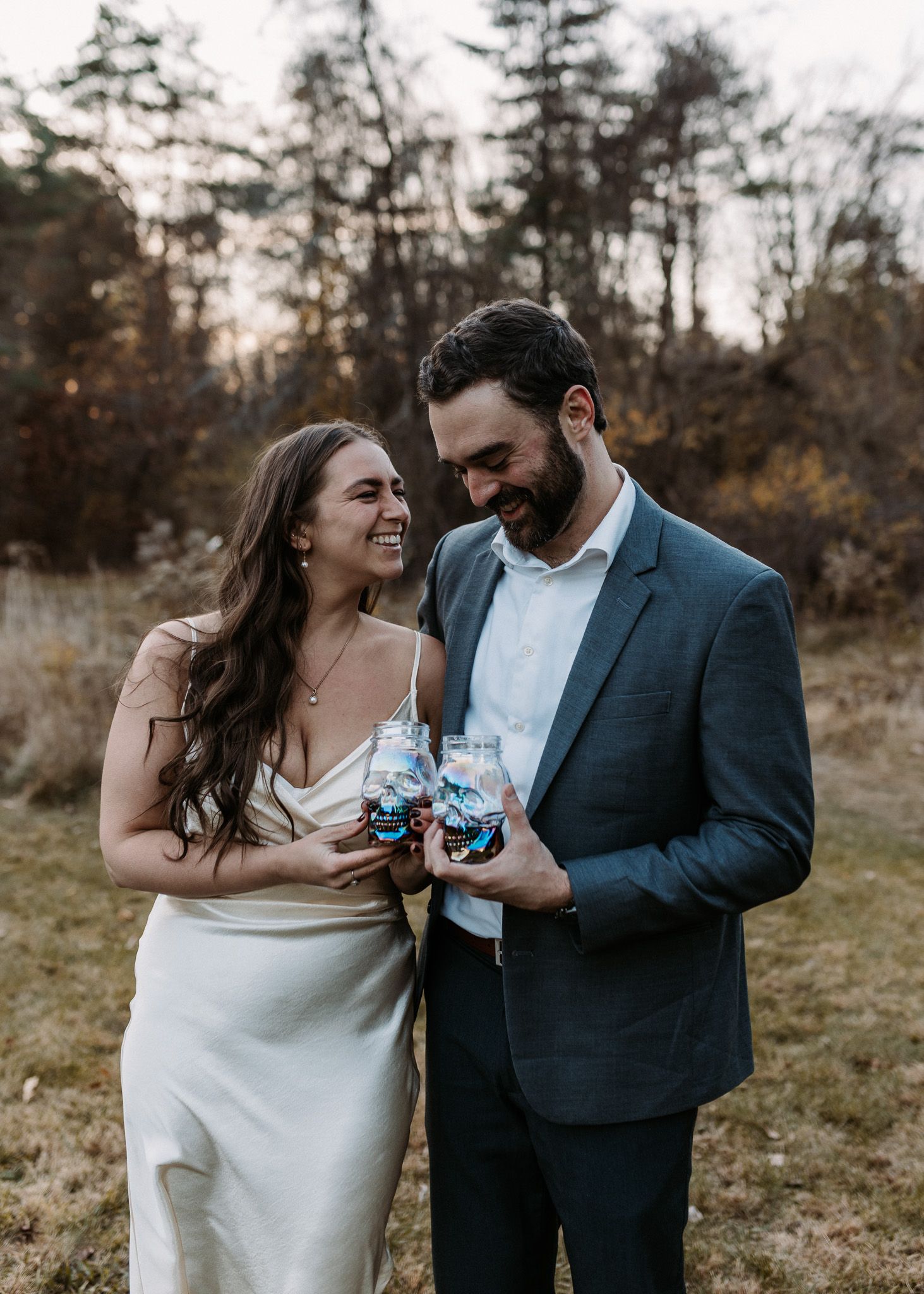 A smiling bride in a white dress and groom in a blue suit hold mason jar drinks outdoors at dusk.