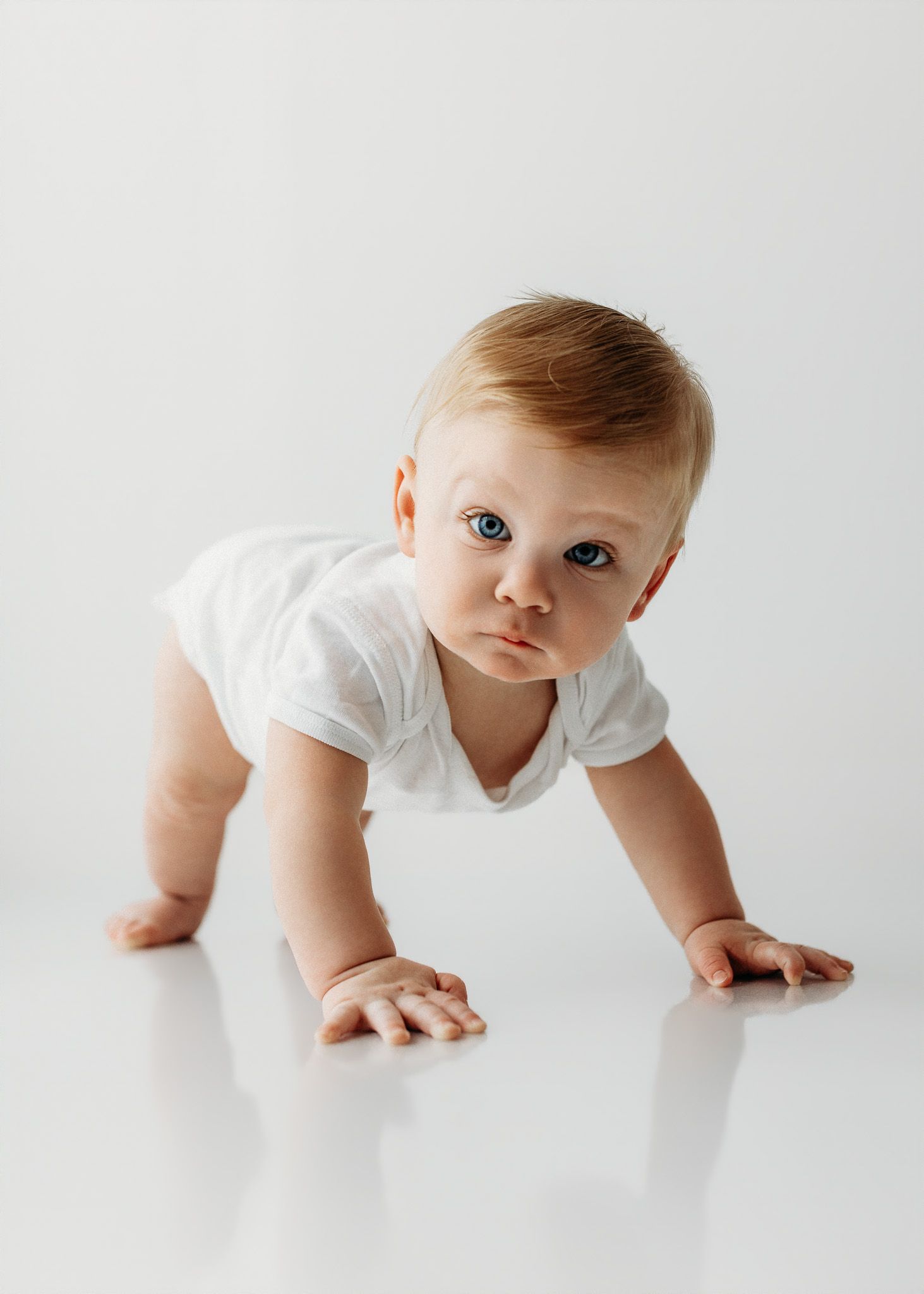 A curious baby in a white onesie crawls on a reflective white floor, looking up with bright blue eyes.