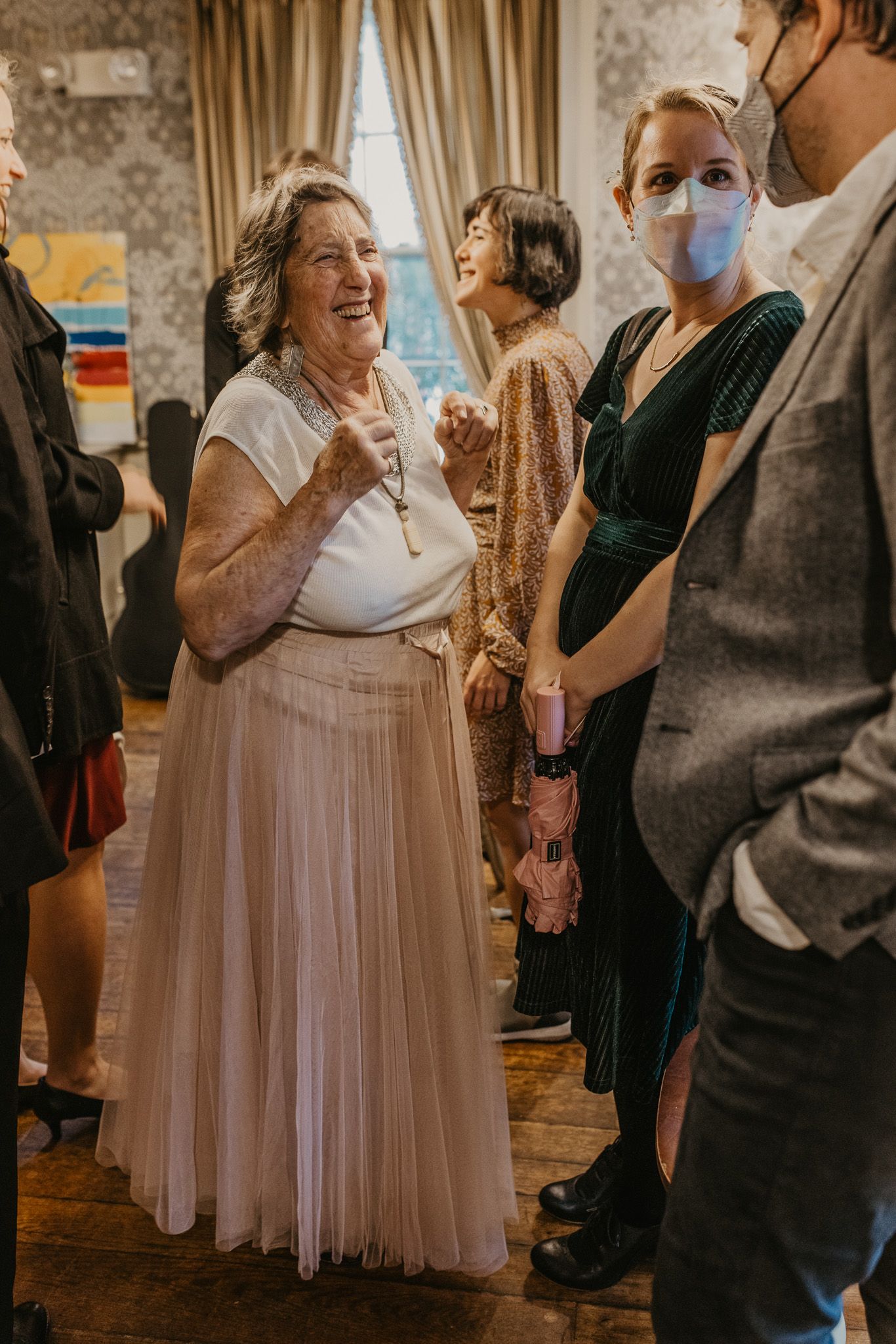 Elderly woman in pink tulle skirt laughing at elegant indoor gathering with guests in formal attire.