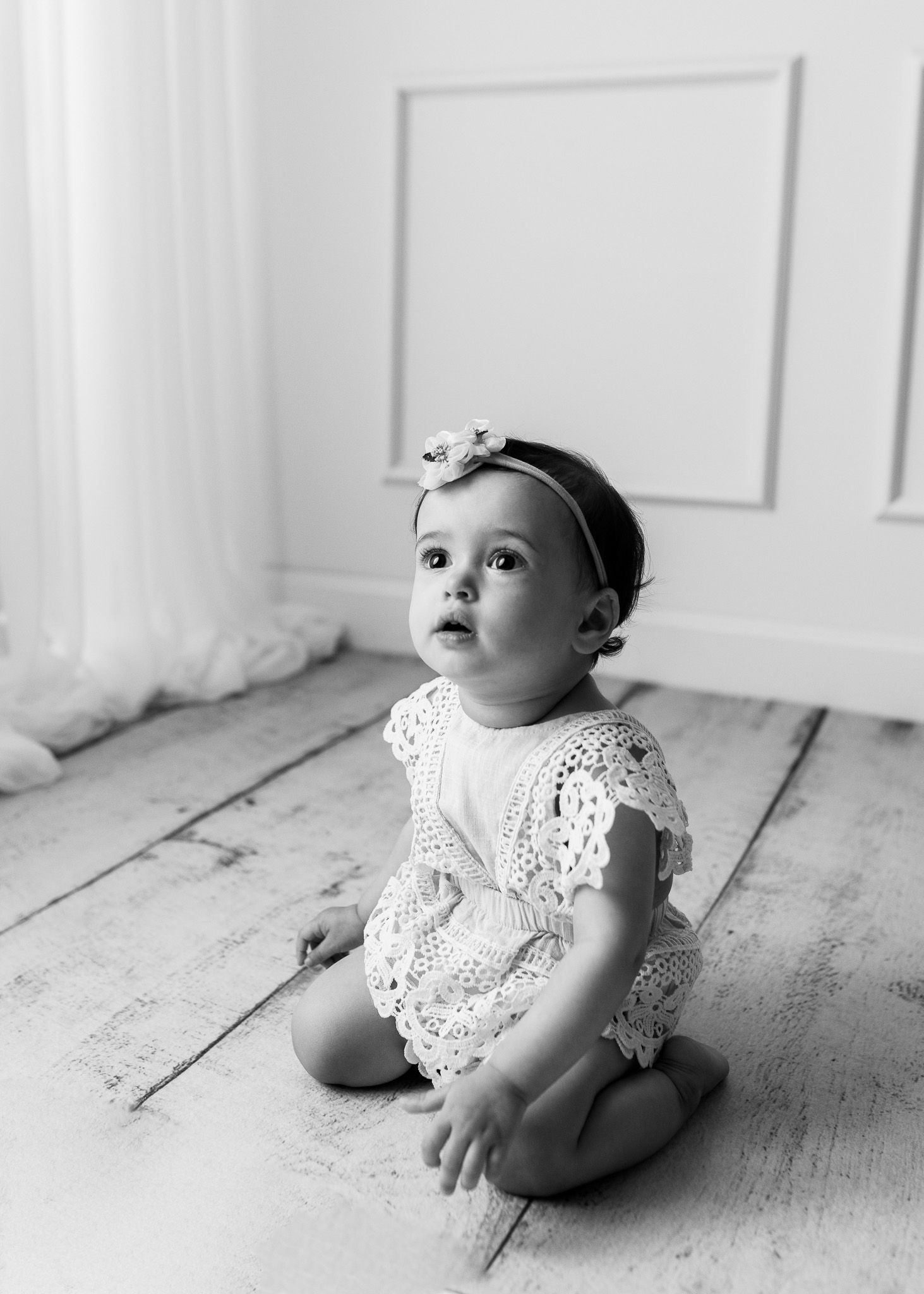 Baby in white lace dress sitting on wooden floor in black and white portrait.