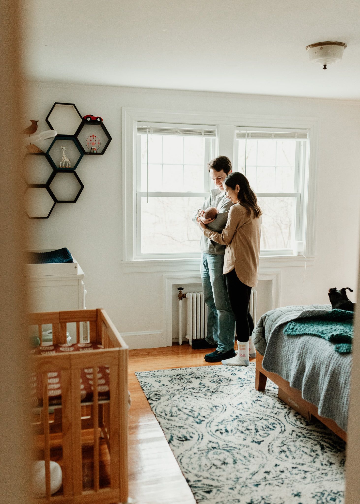 A couple stands by a bright window in a nursery, lovingly holding their newborn baby near a wooden crib.