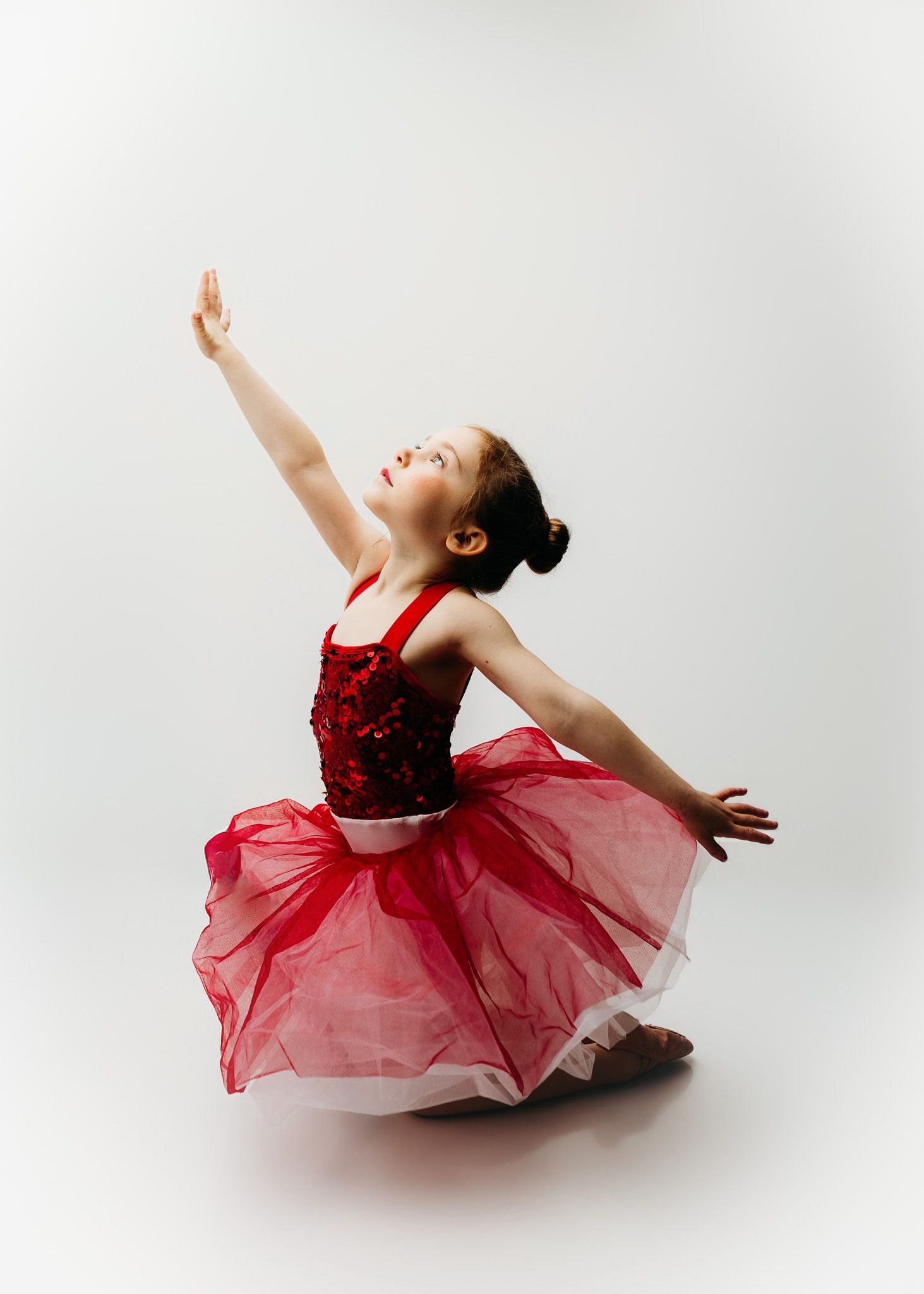 Young girl in red sequin ballet tutu performs graceful dance pose with arm raised against white background.