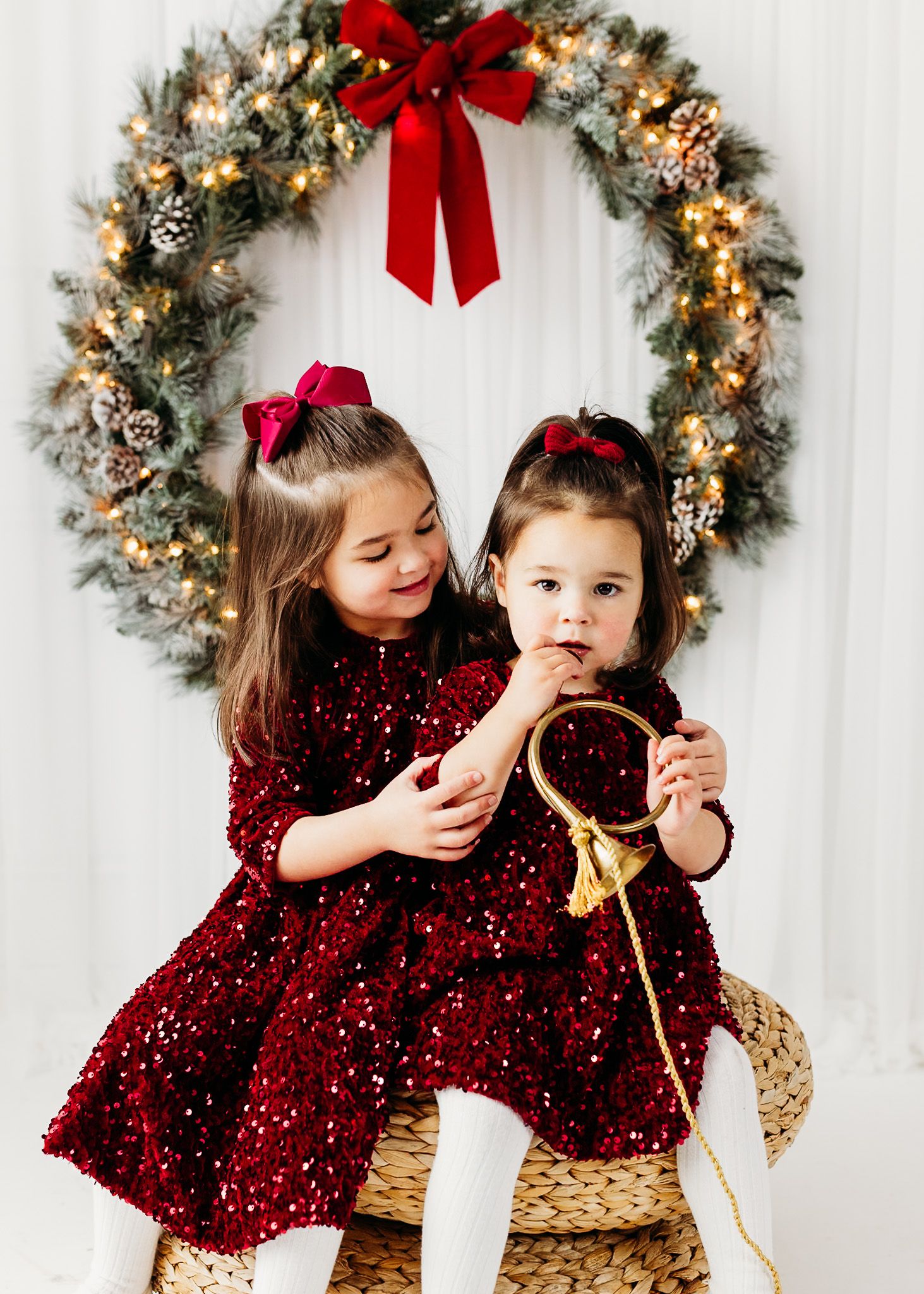 Two young girls in matching red sequin dresses pose before a lit Christmas wreath with a red bow.