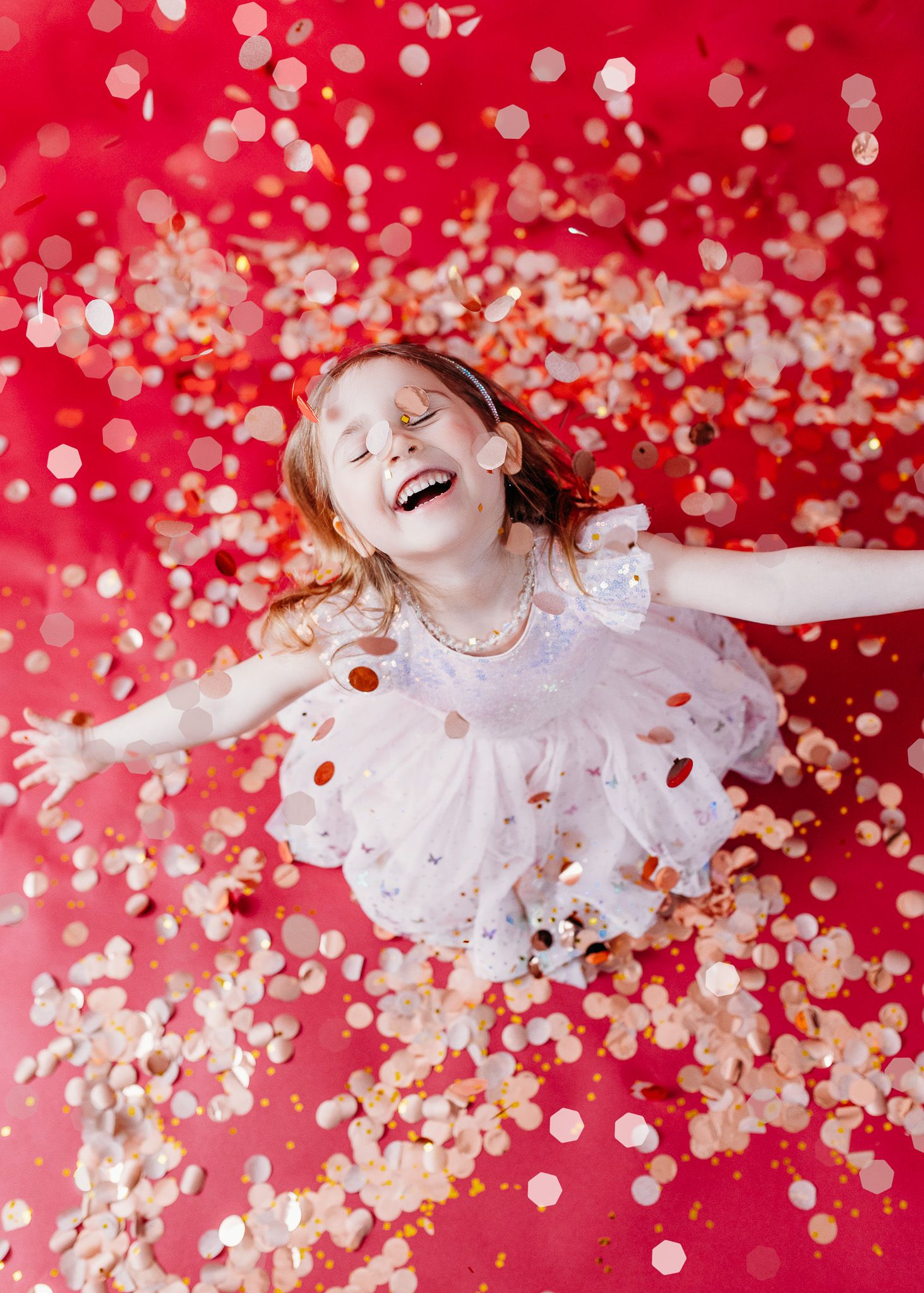 Child in white dress lying among gold confetti on red background.