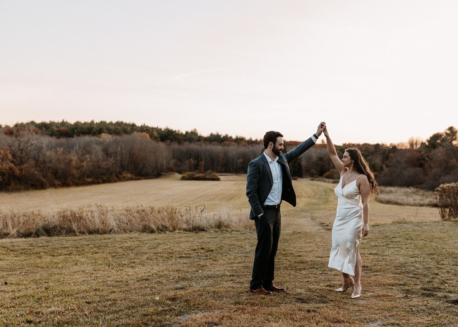 Couple dancing outdoors at golden hour in a field, groom in dark suit, bride in white gown, autumn trees backdrop.