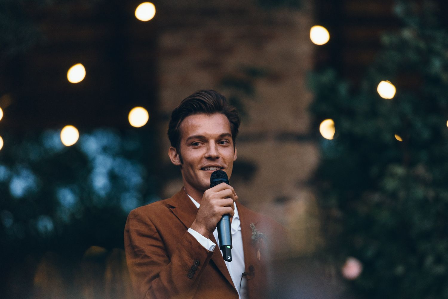 A speaker holds a microphone at an evening event with string lights in the background.