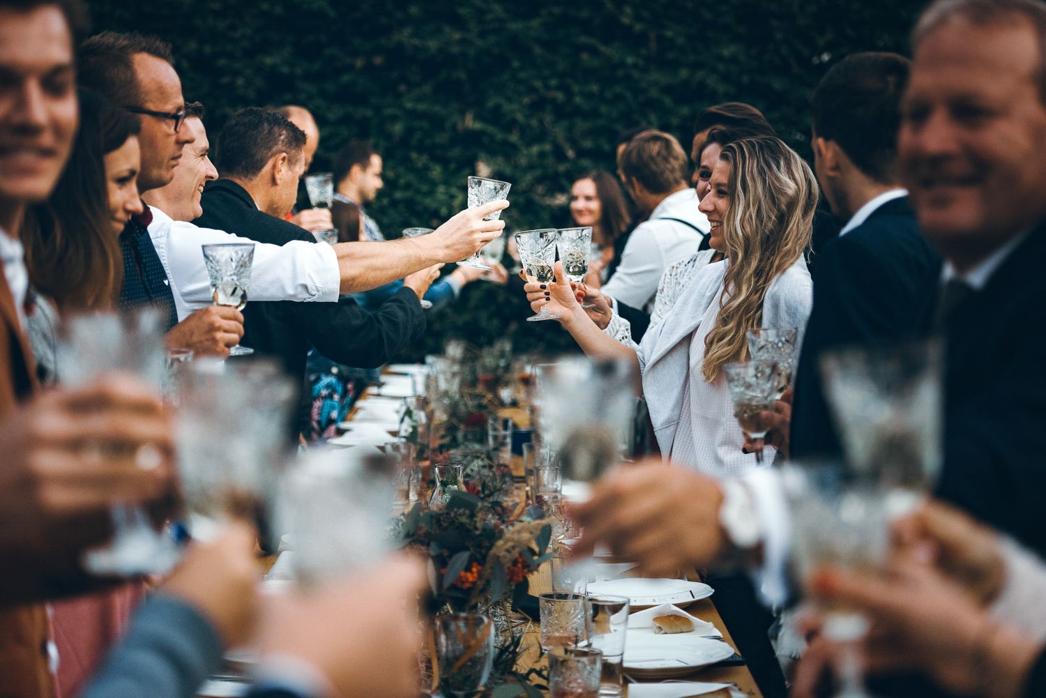 Wedding guests toasting with champagne glasses at a long outdoor reception table.