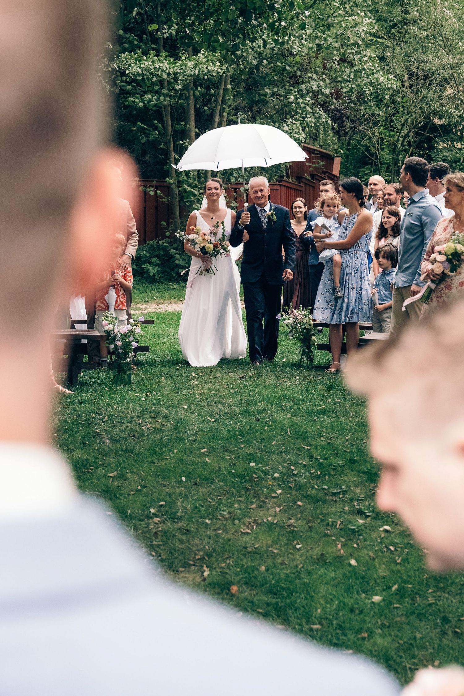 Wedding guests gathered on a green lawn with white umbrellas during an outdoor ceremony.