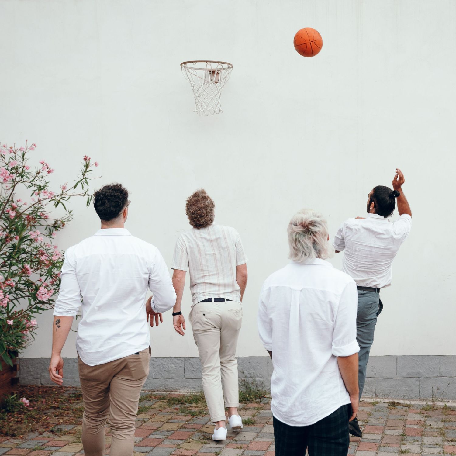 Four people in white shirts playing basketball outdoors near flowering bushes.