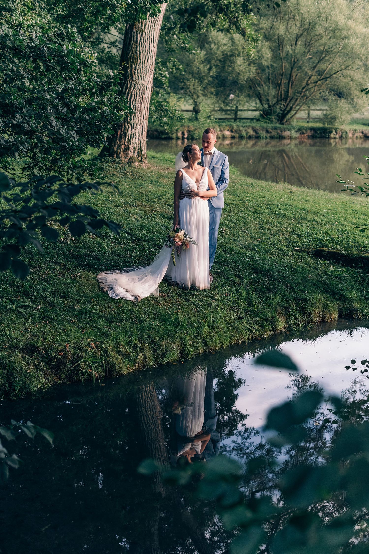 Romantic wedding couple poses by a reflective pond surrounded by trees in a serene outdoor setting.