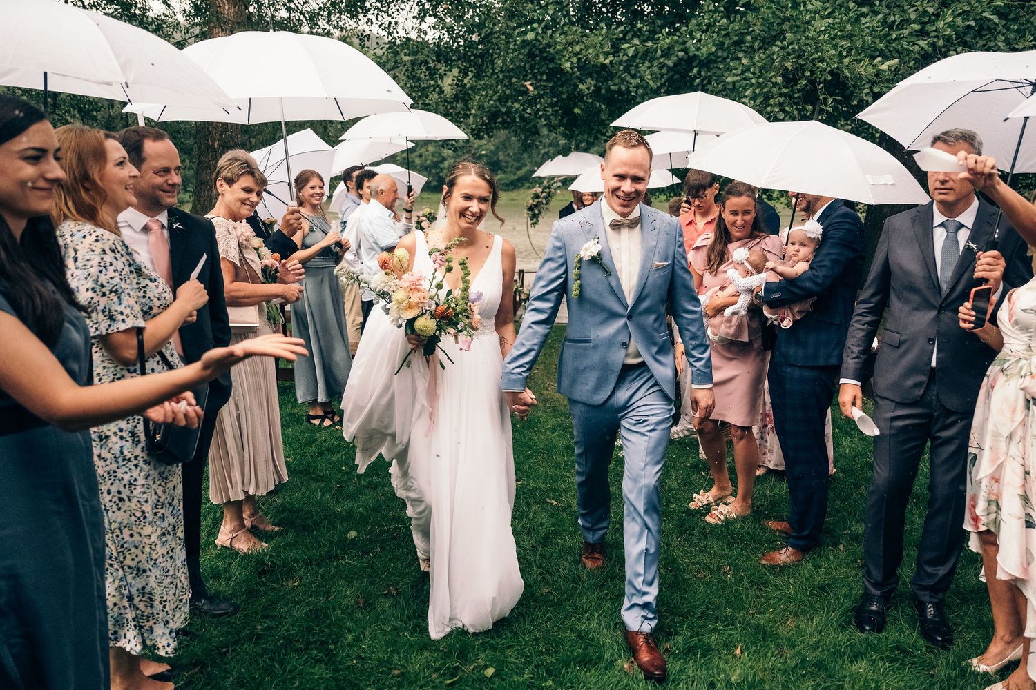 Wedding guests with umbrellas celebrate during outdoor ceremony near a lake.