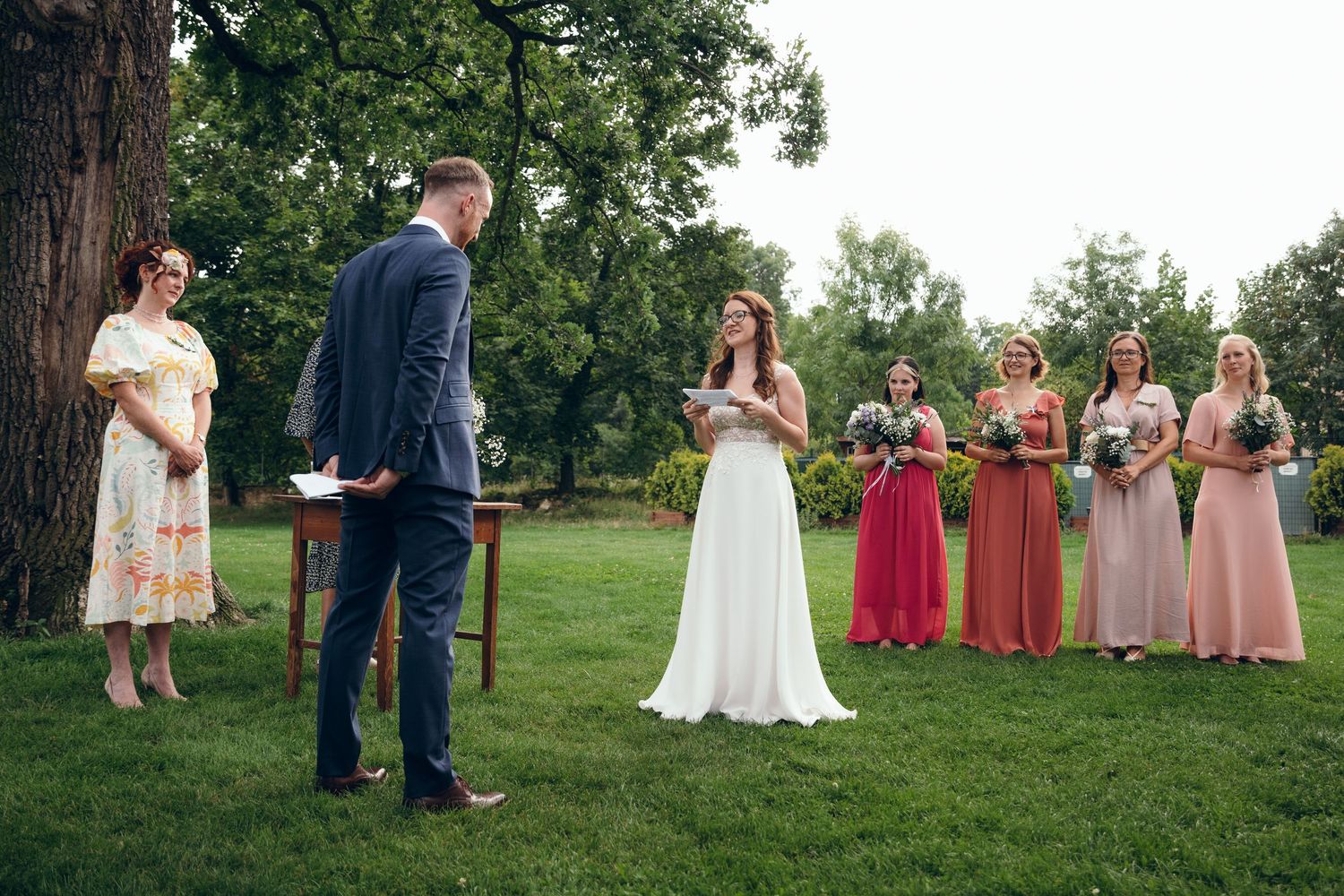 Wedding party poses on green lawn with bridesmaids in coral dresses.