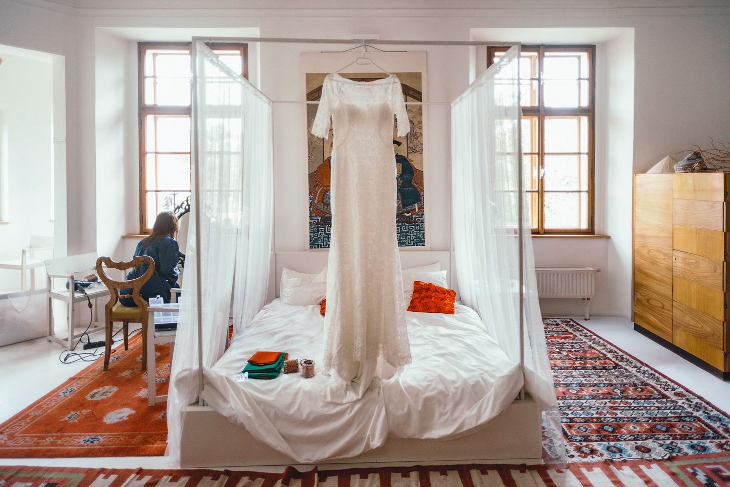 An elegant white wedding dress displayed on a bed in a bright room with large windows.