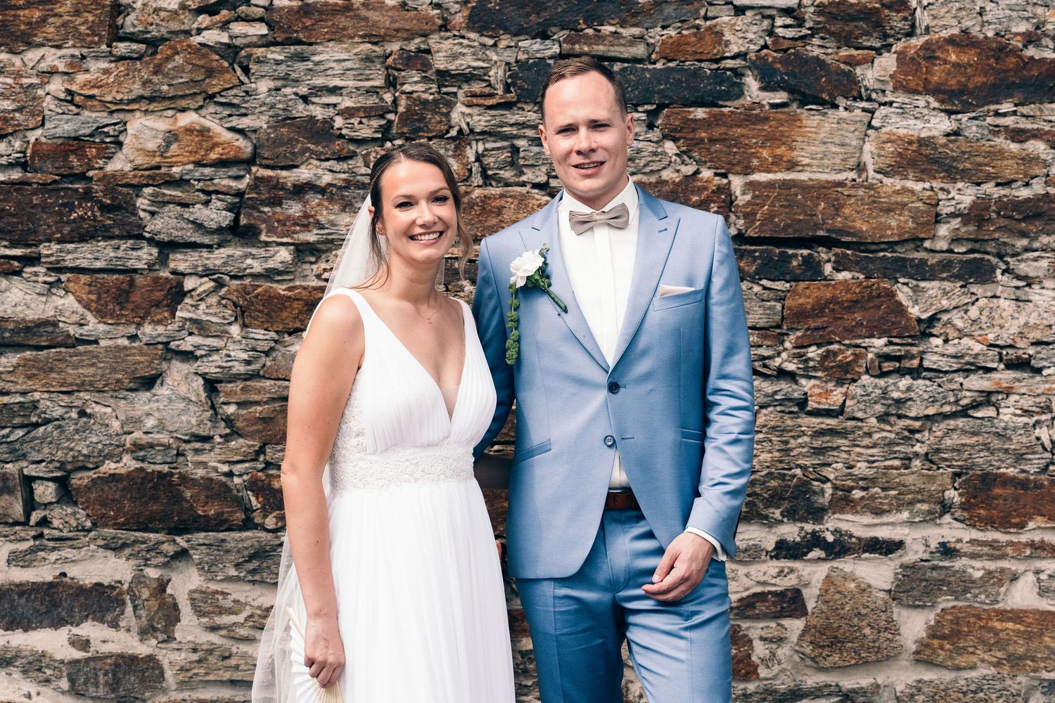Wedding couple poses against rustic stone wall backdrop in elegant attire.
