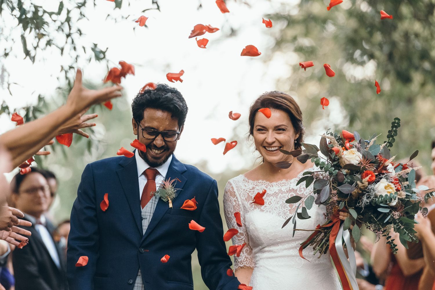 Red rose petals shower down during a joyful outdoor wedding celebration.
