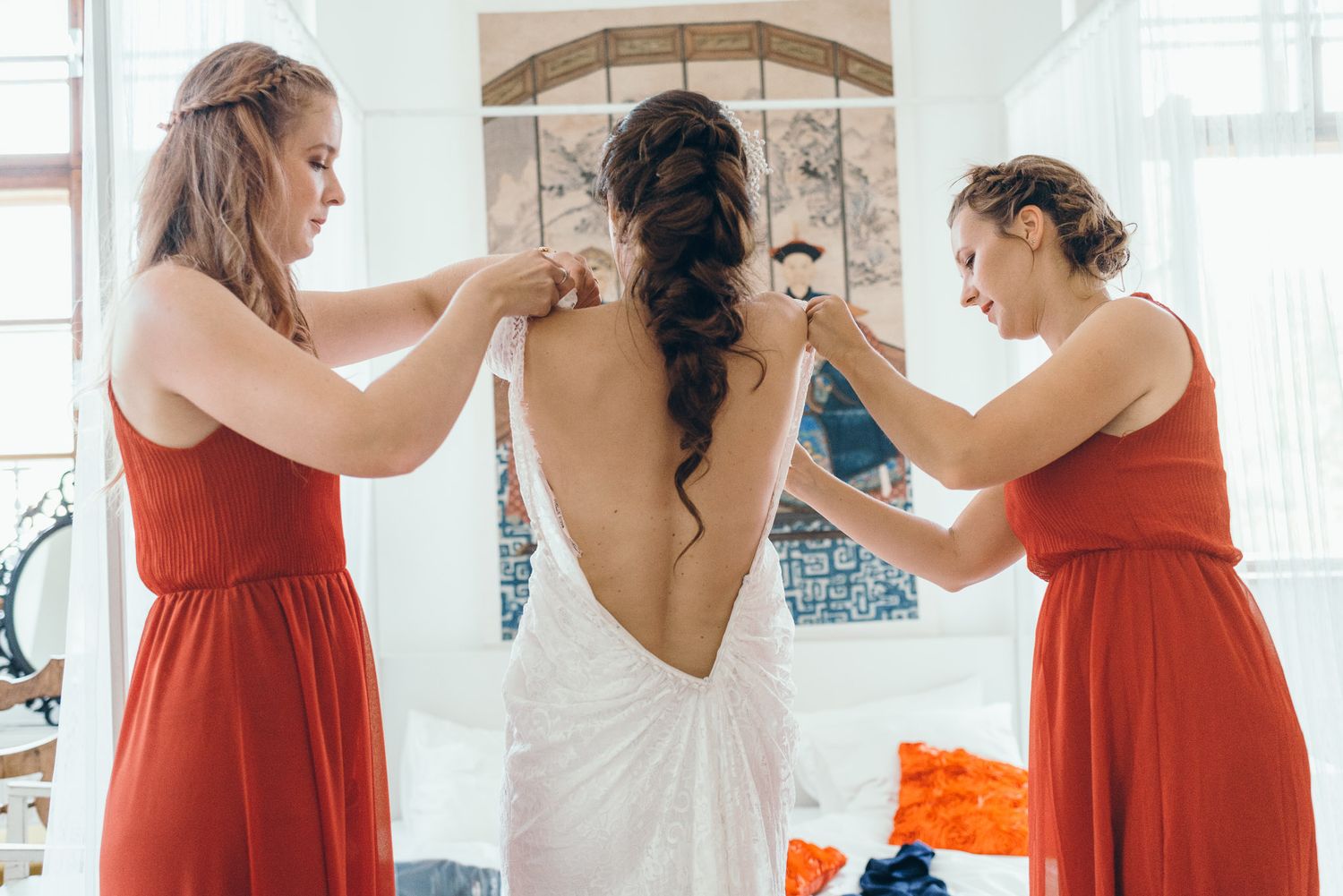 Bridesmaids in red dresses help with wedding preparations in a bright white room.