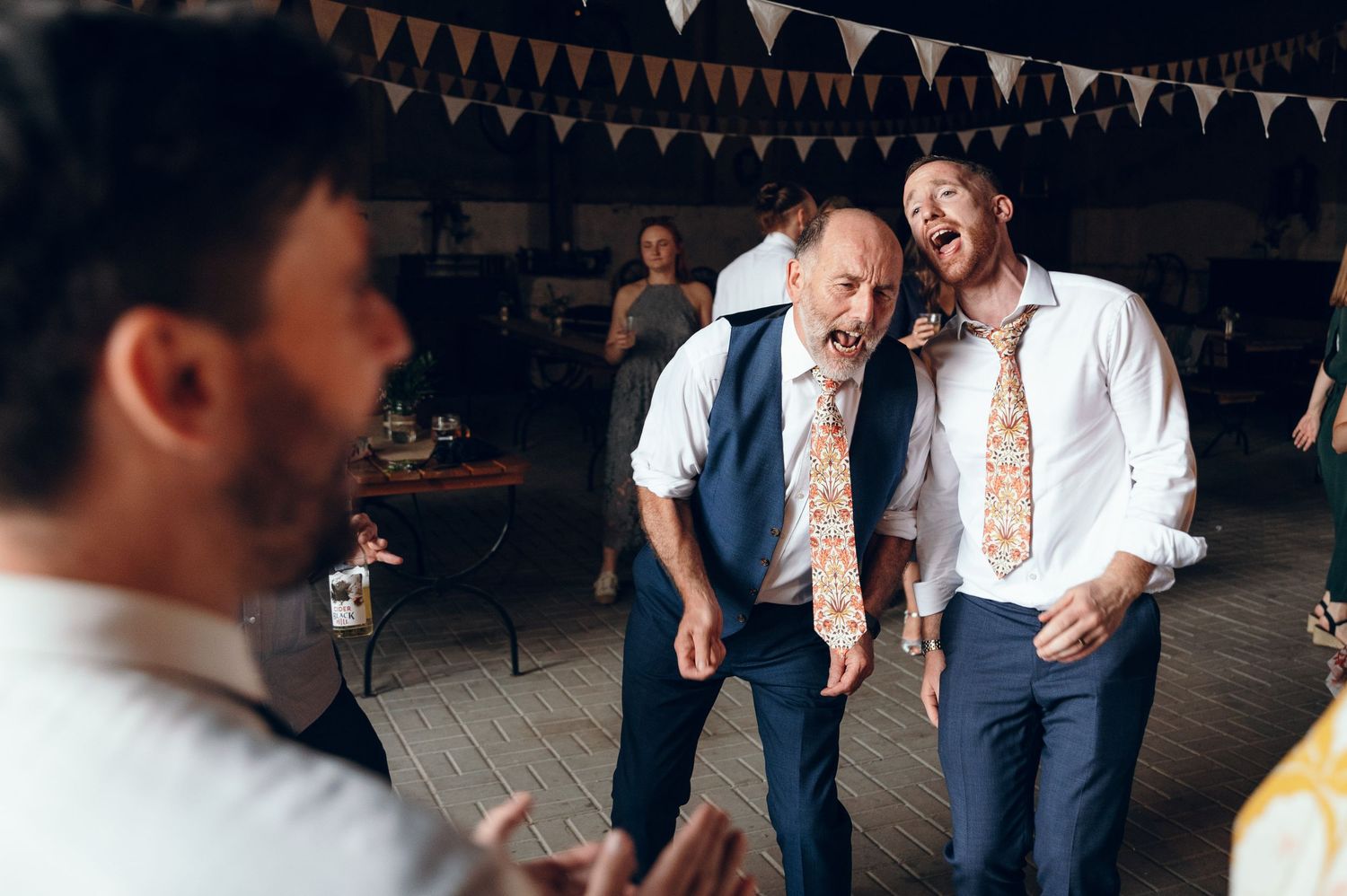 Party guests in white shirts and ties dancing enthusiastically under festive bunting decorations.