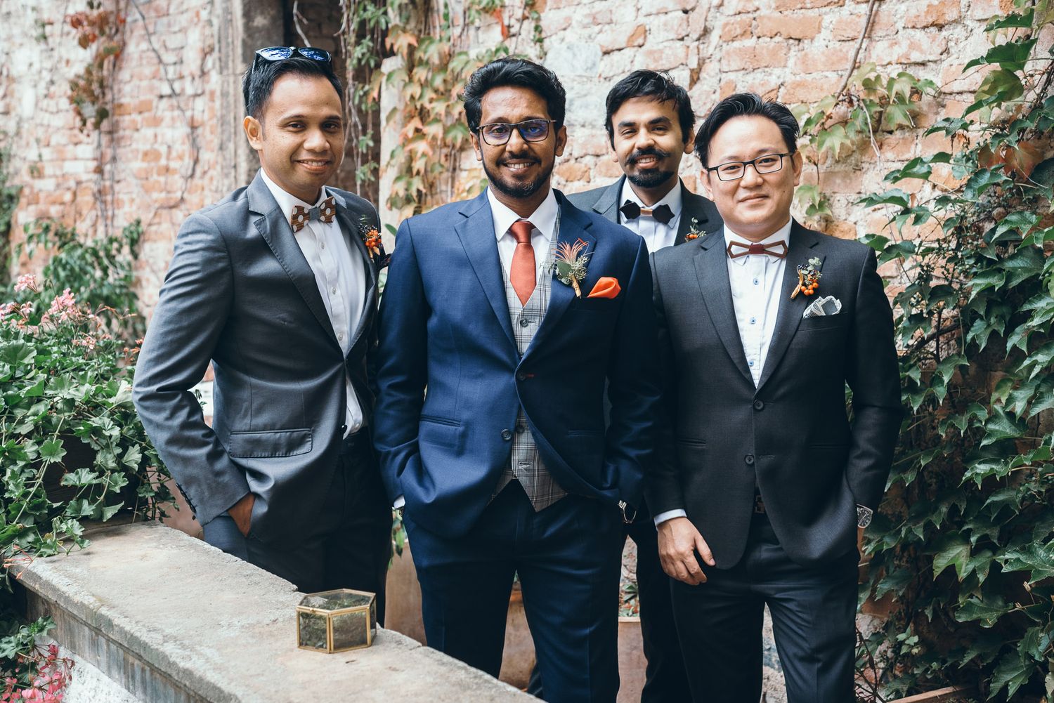 Groomsmen in dark suits pose together against a rustic brick wall backdrop at a wedding celebration.