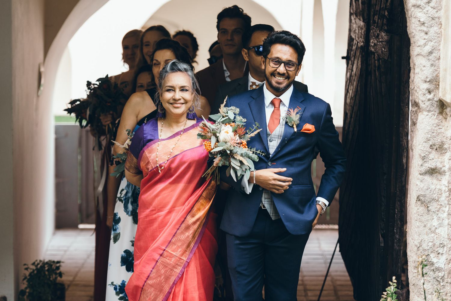 Wedding party members celebrate during an outdoor ceremony under white arches.