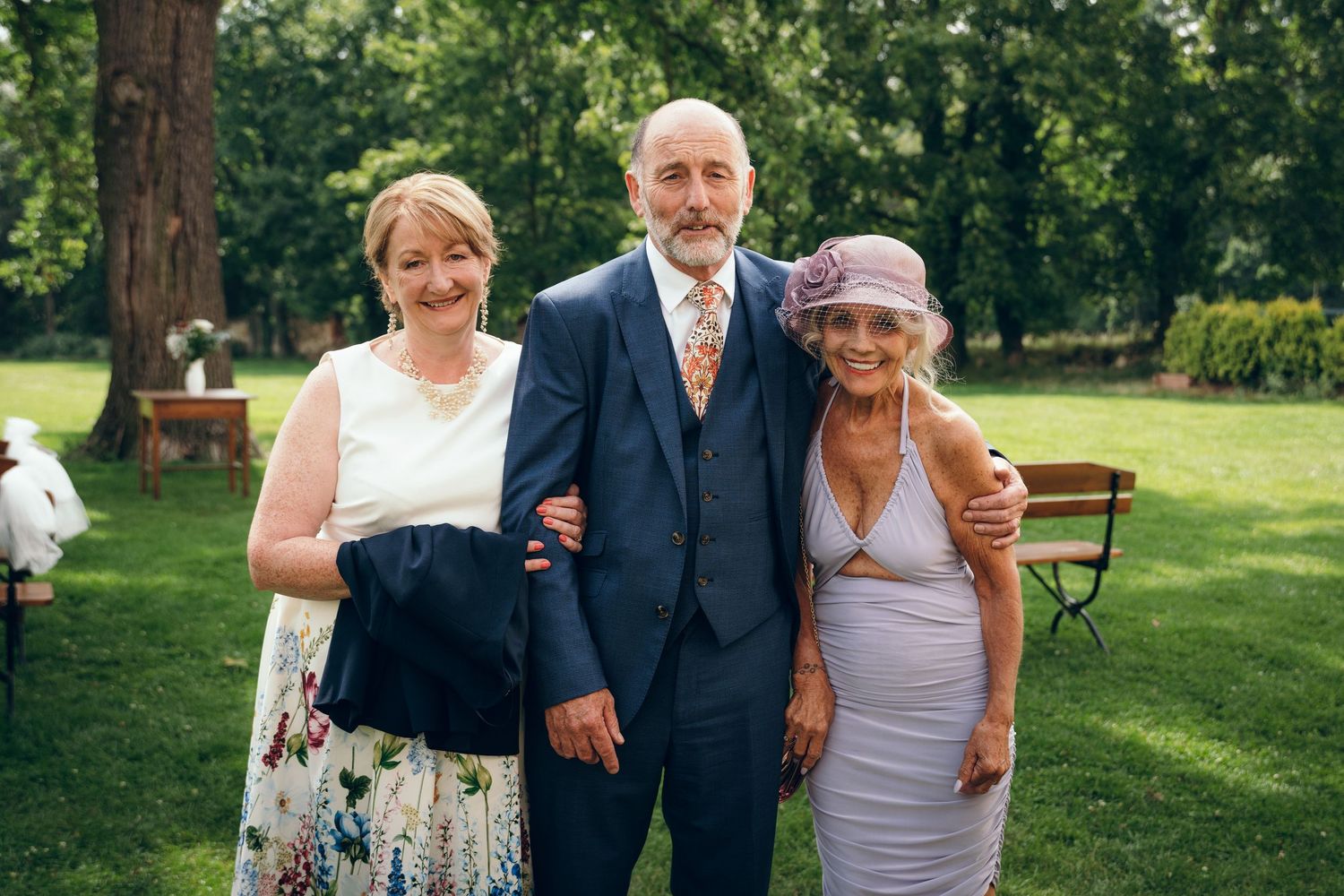 Wedding party members gather for photos on a sunny lawn with trees in the background.