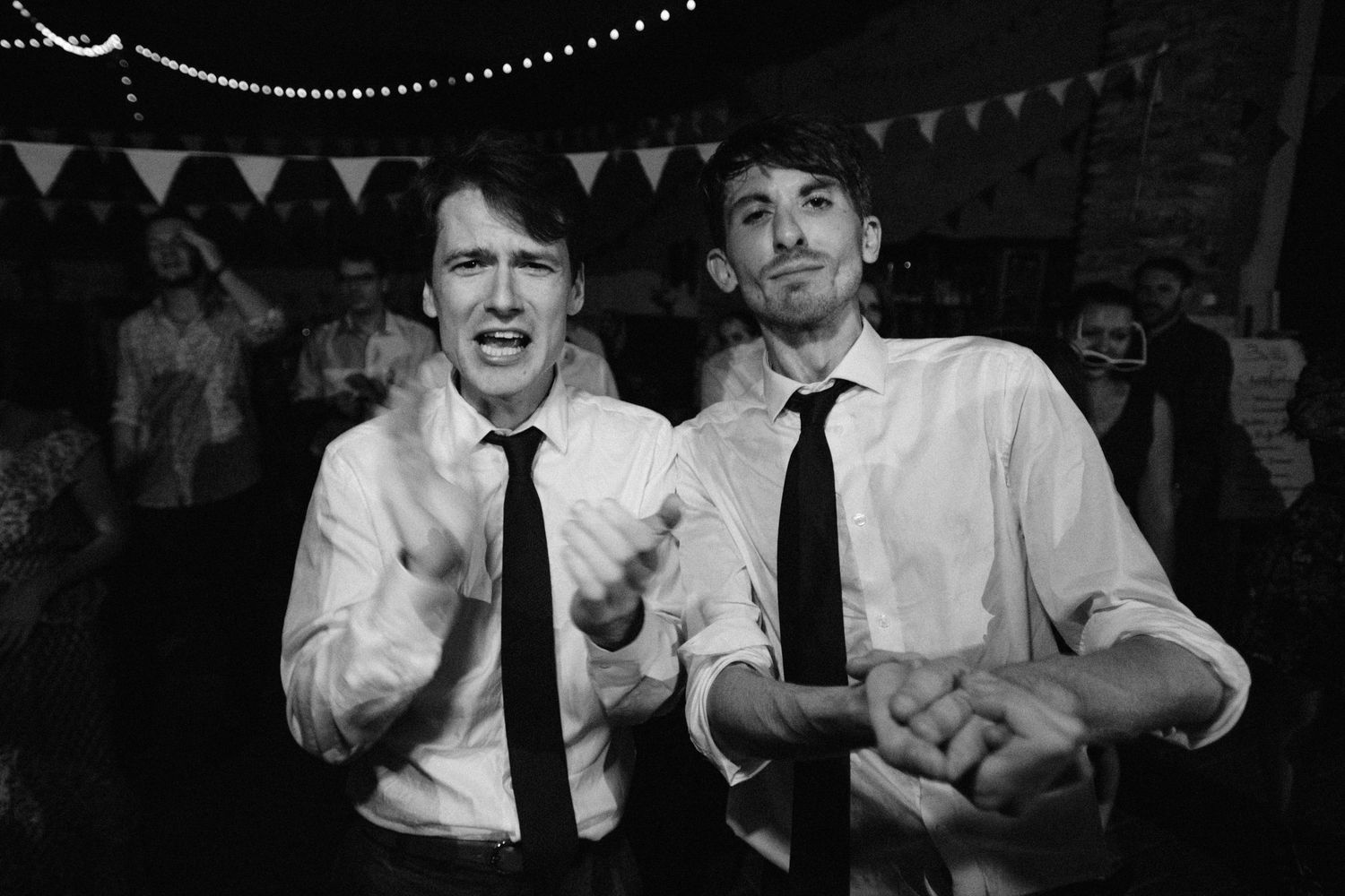 Two men in white shirts and black ties laughing together at a party with string lights and bunting in the background.