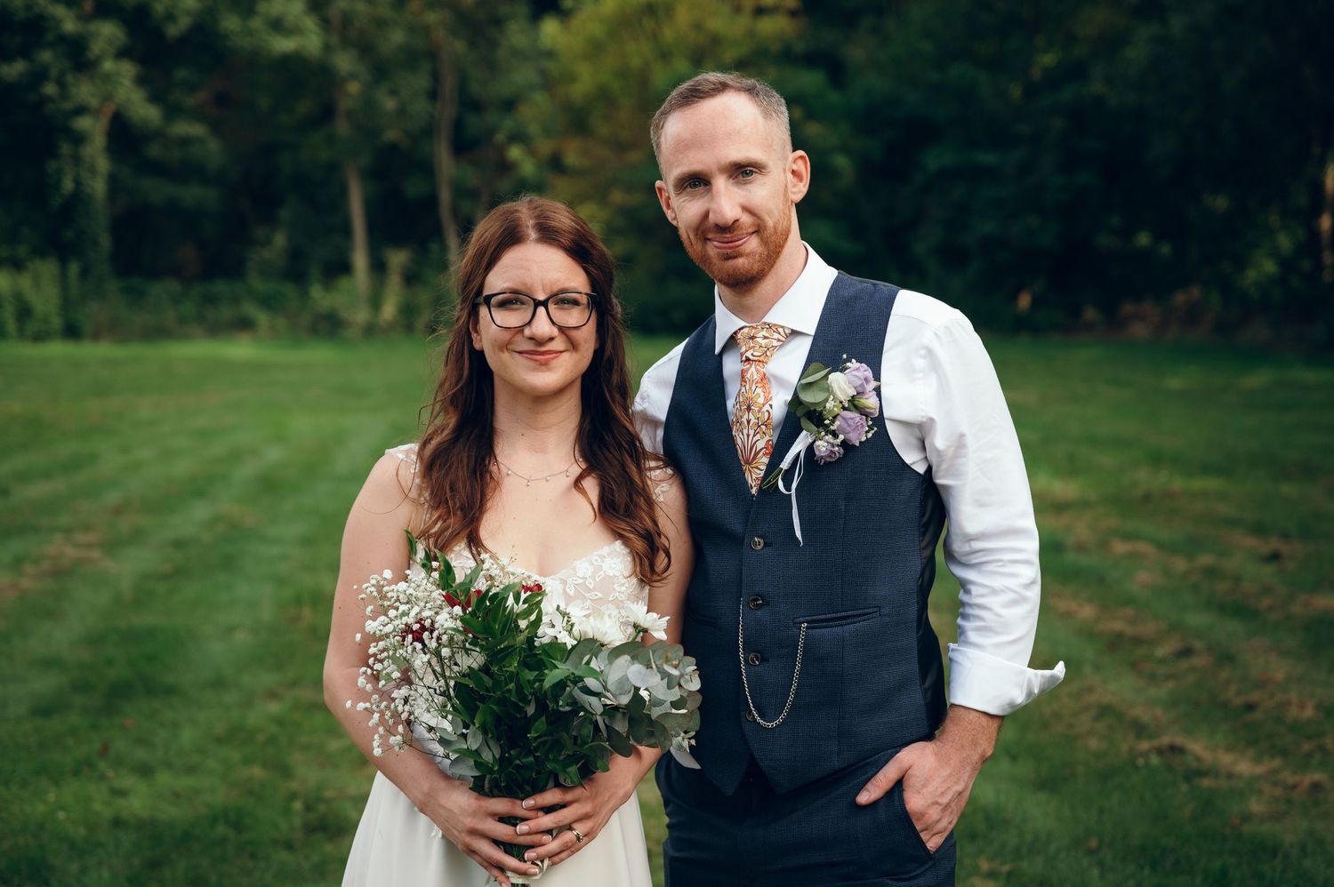 A wedding couple poses outdoors with greenery in the background, holding bouquets.