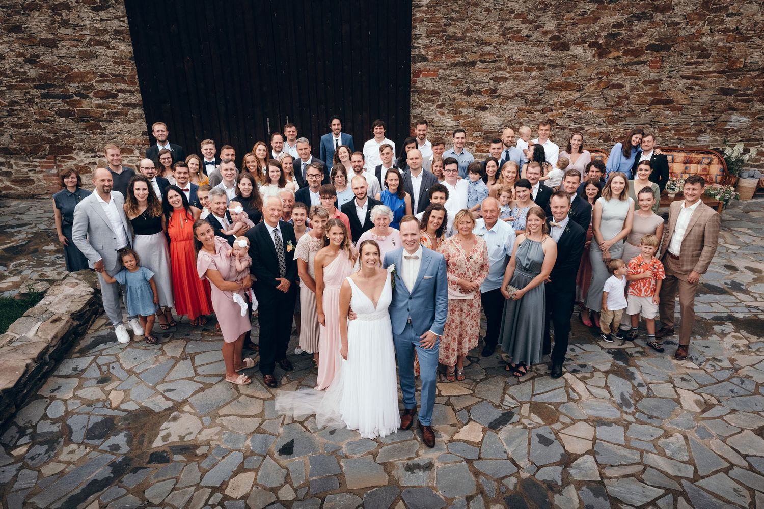Large group wedding photo of all guests against rustic stone wall background.