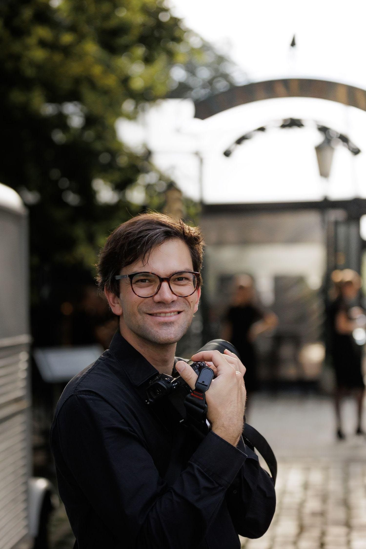 A person in glasses and dark clothing smiles while posing outdoors near a white architectural structure.