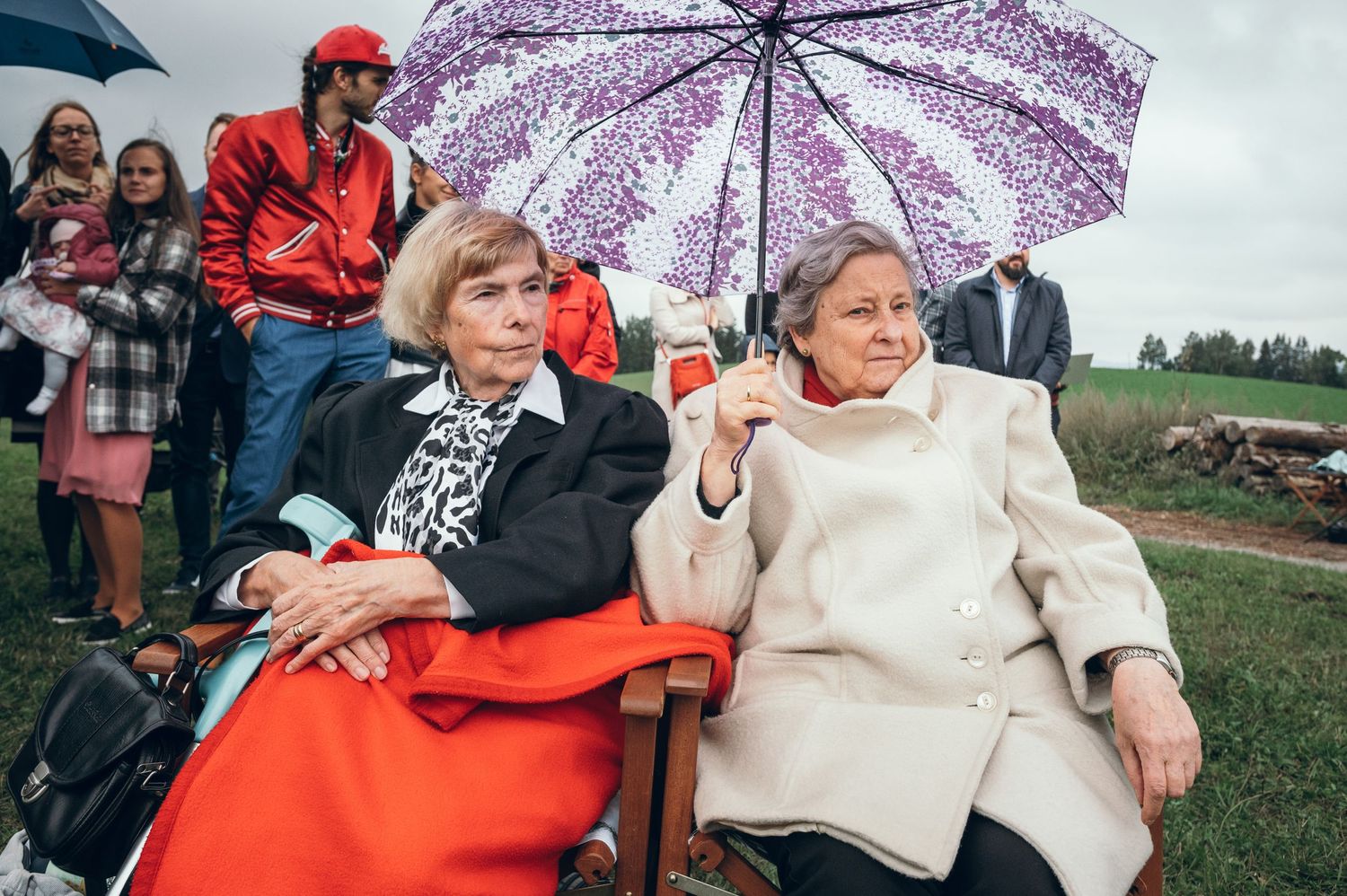 Two people sitting in chairs outdoors, one with a floral umbrella and wearing bright colors.