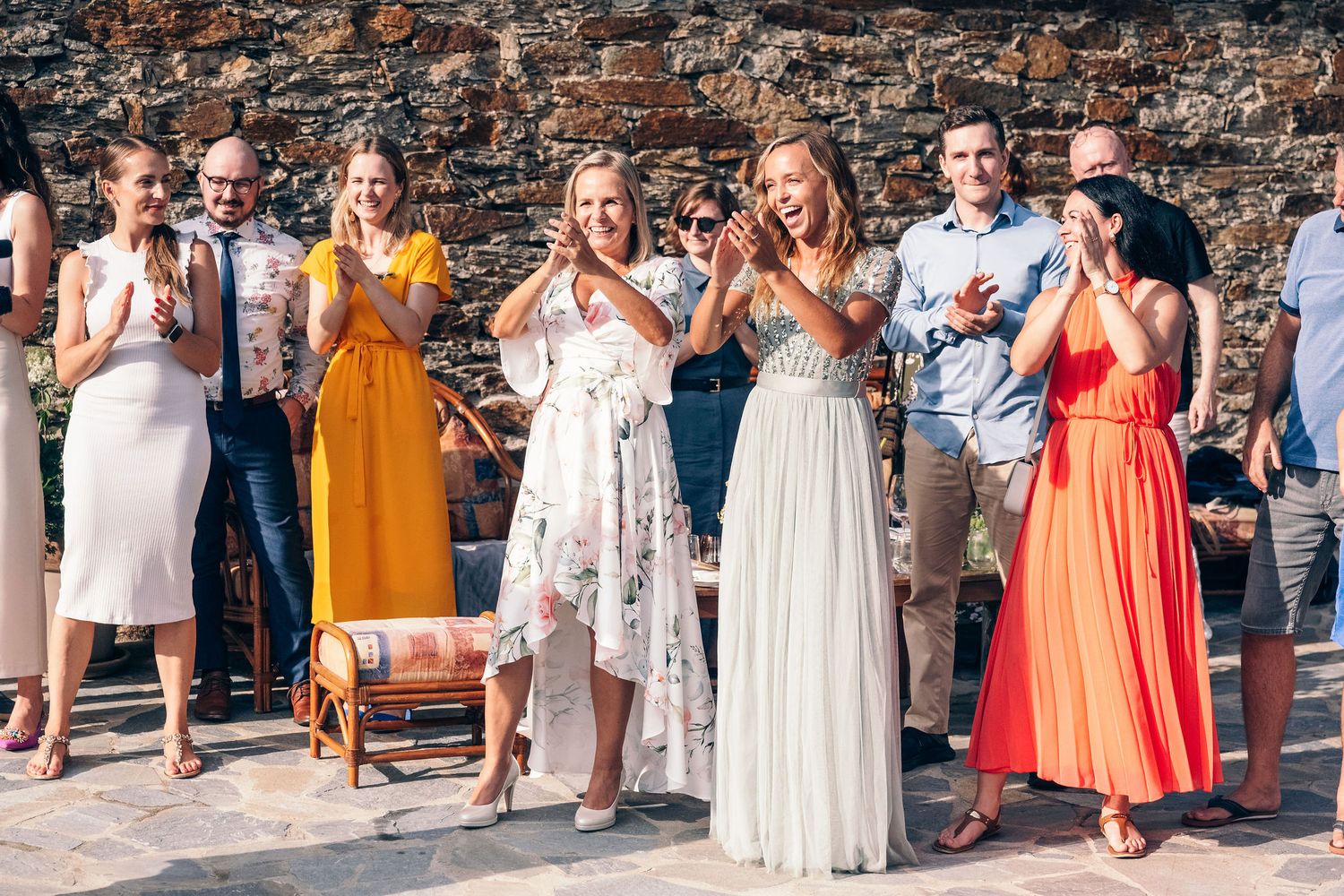 Wedding guests in colorful dresses line up against stone wall for group photo.