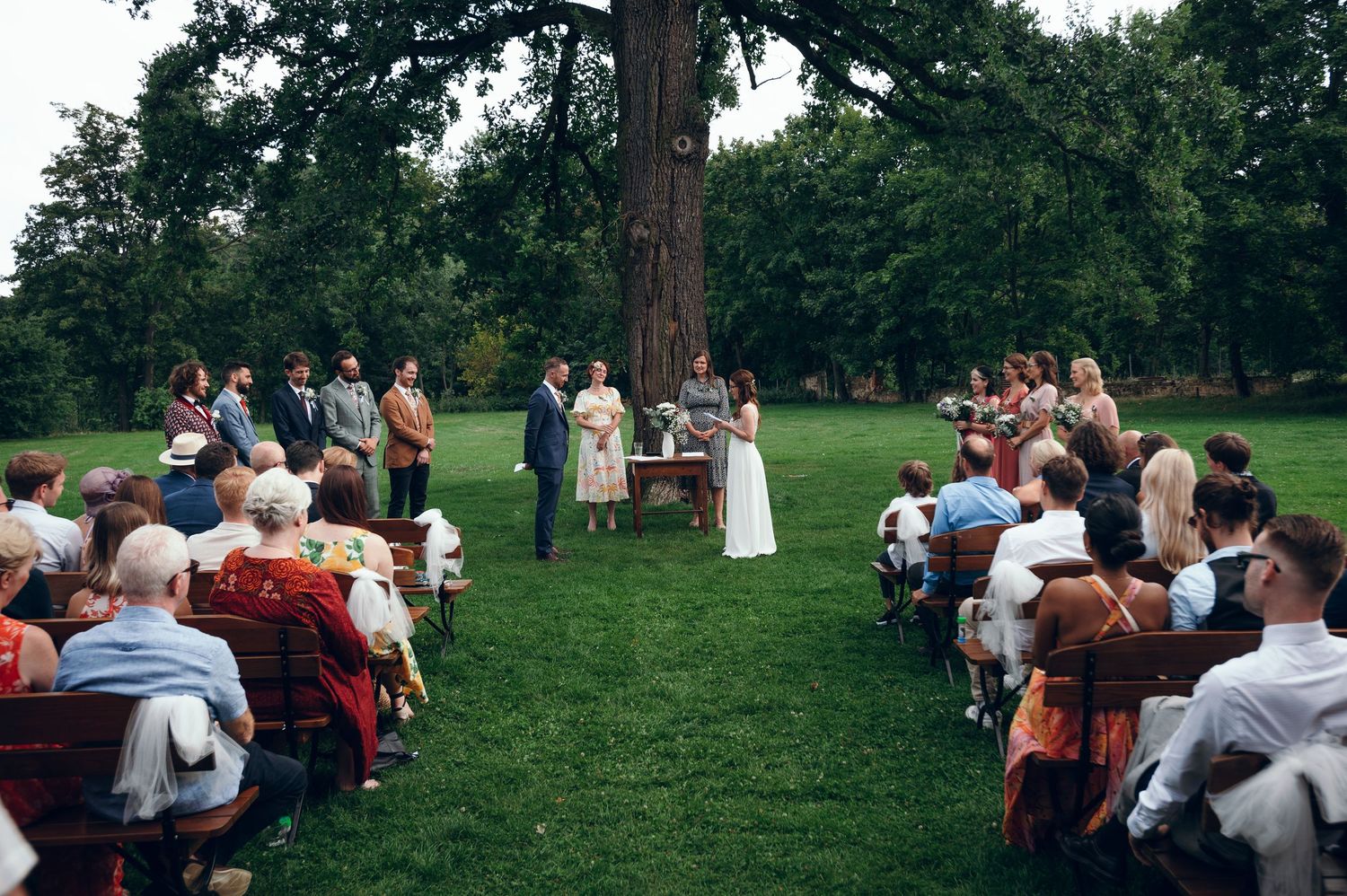 Wedding guests seated on wooden chairs during outdoor ceremony among trees.