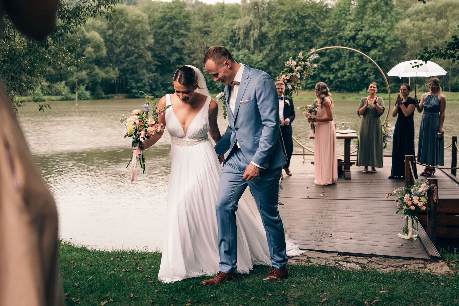Scenic outdoor wedding ceremony by a lake with guests seated on wooden chairs.