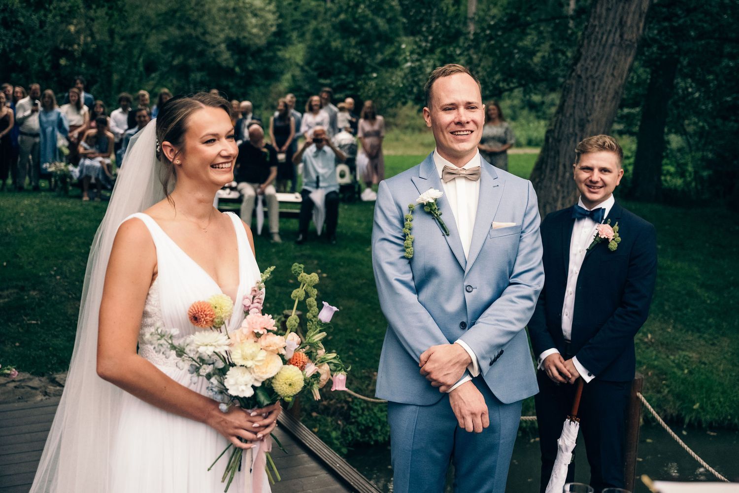 Wedding party in light gray suits and white dresses gather outdoors for group photos.