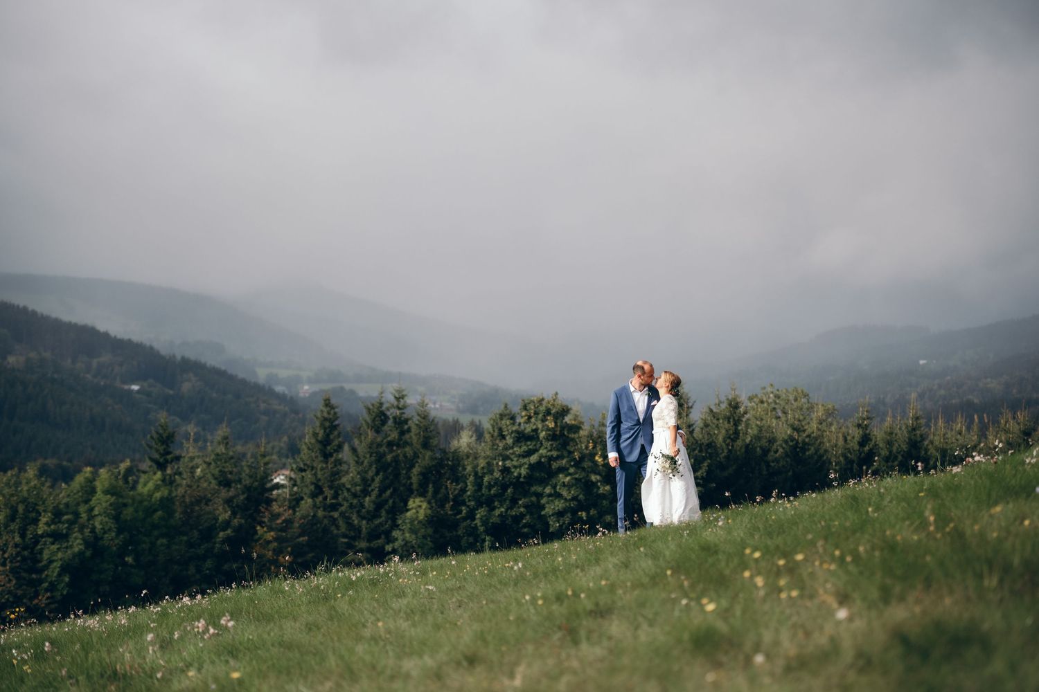 Newlyweds standing on a hilltop meadow with misty mountains in the background.