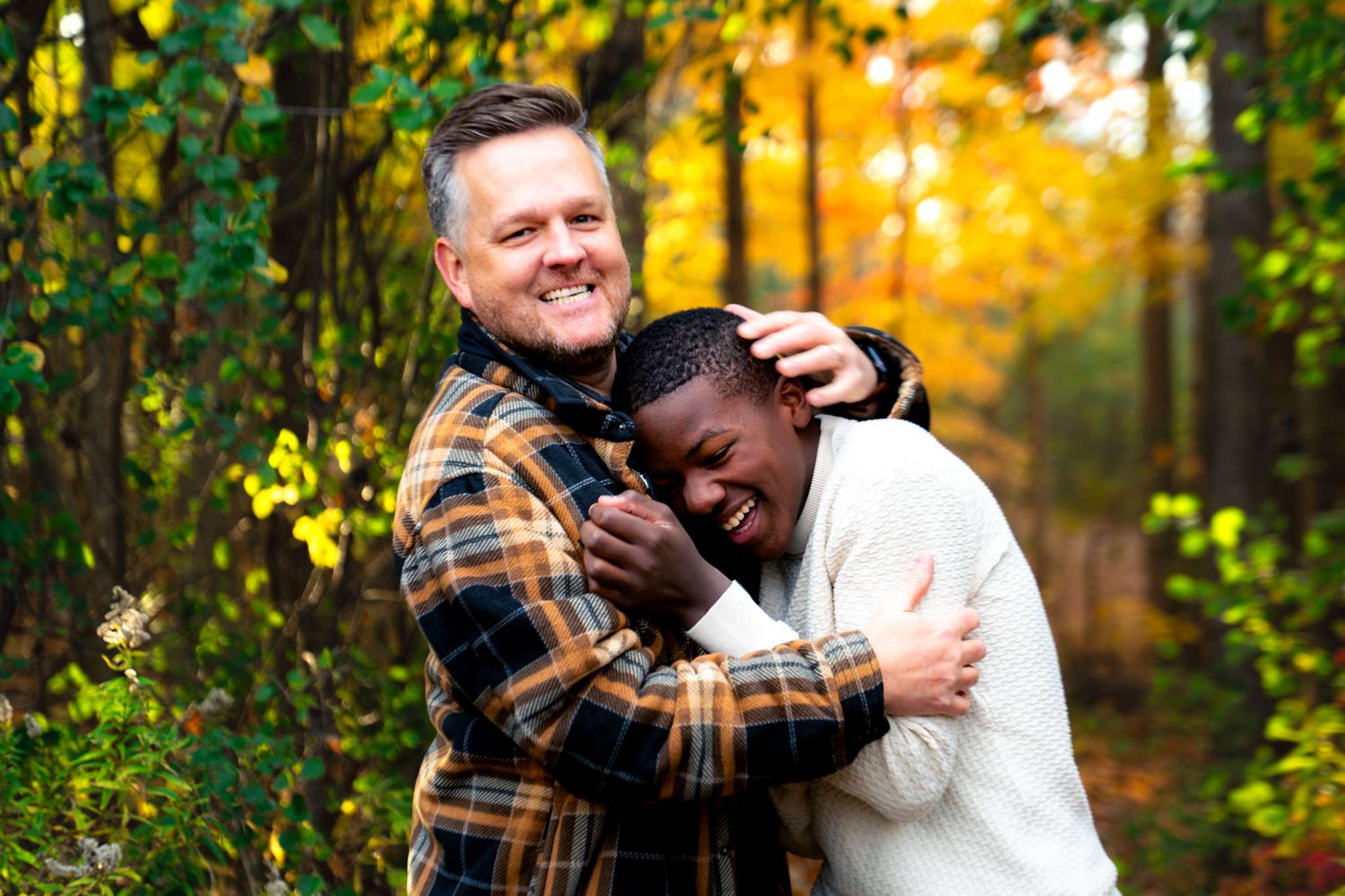 Two people share a warm embrace outdoors during golden hour with autumn foliage in the background.