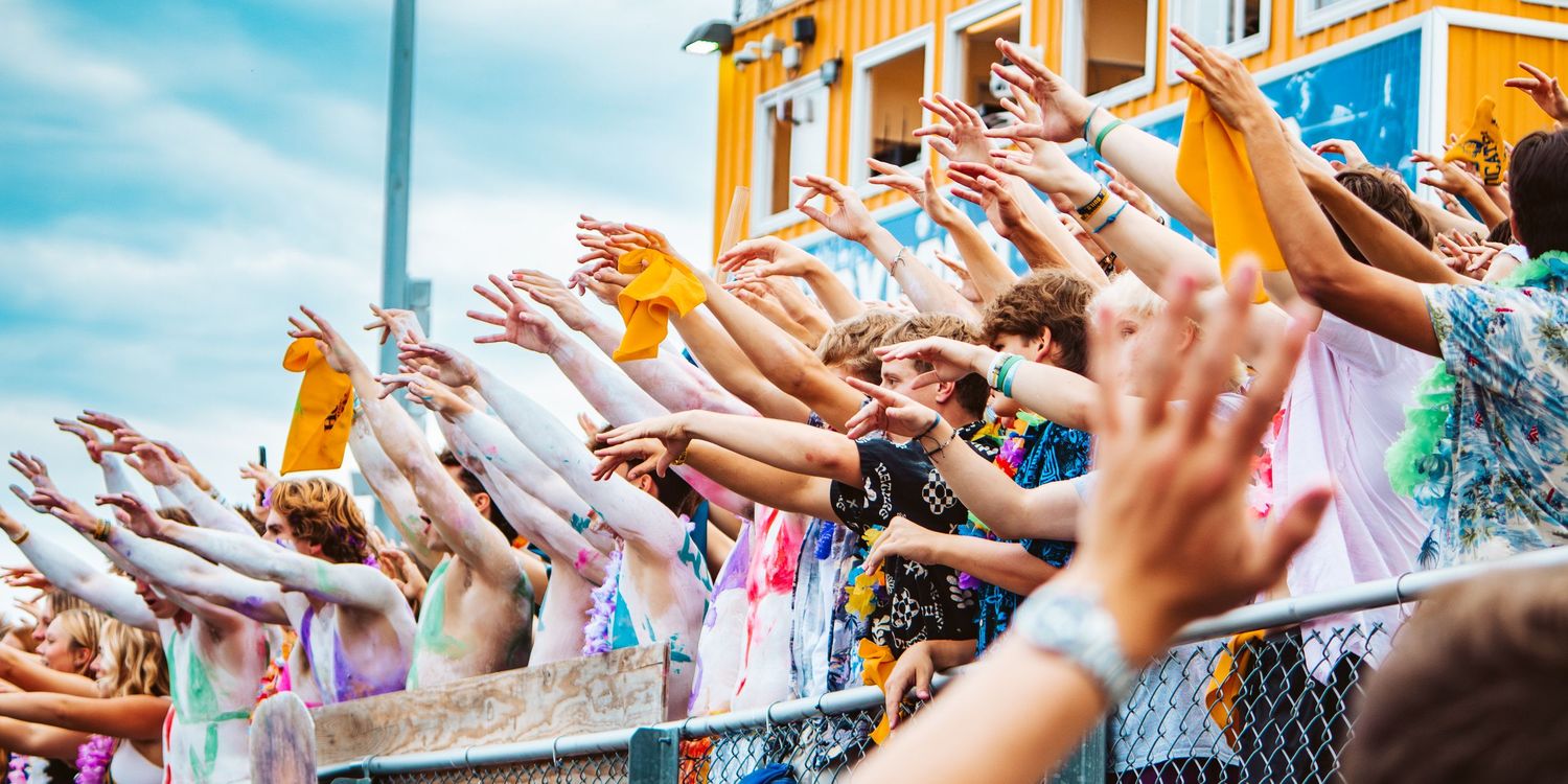 Row of raised hands reaching upward against bright blue sky and orange building.