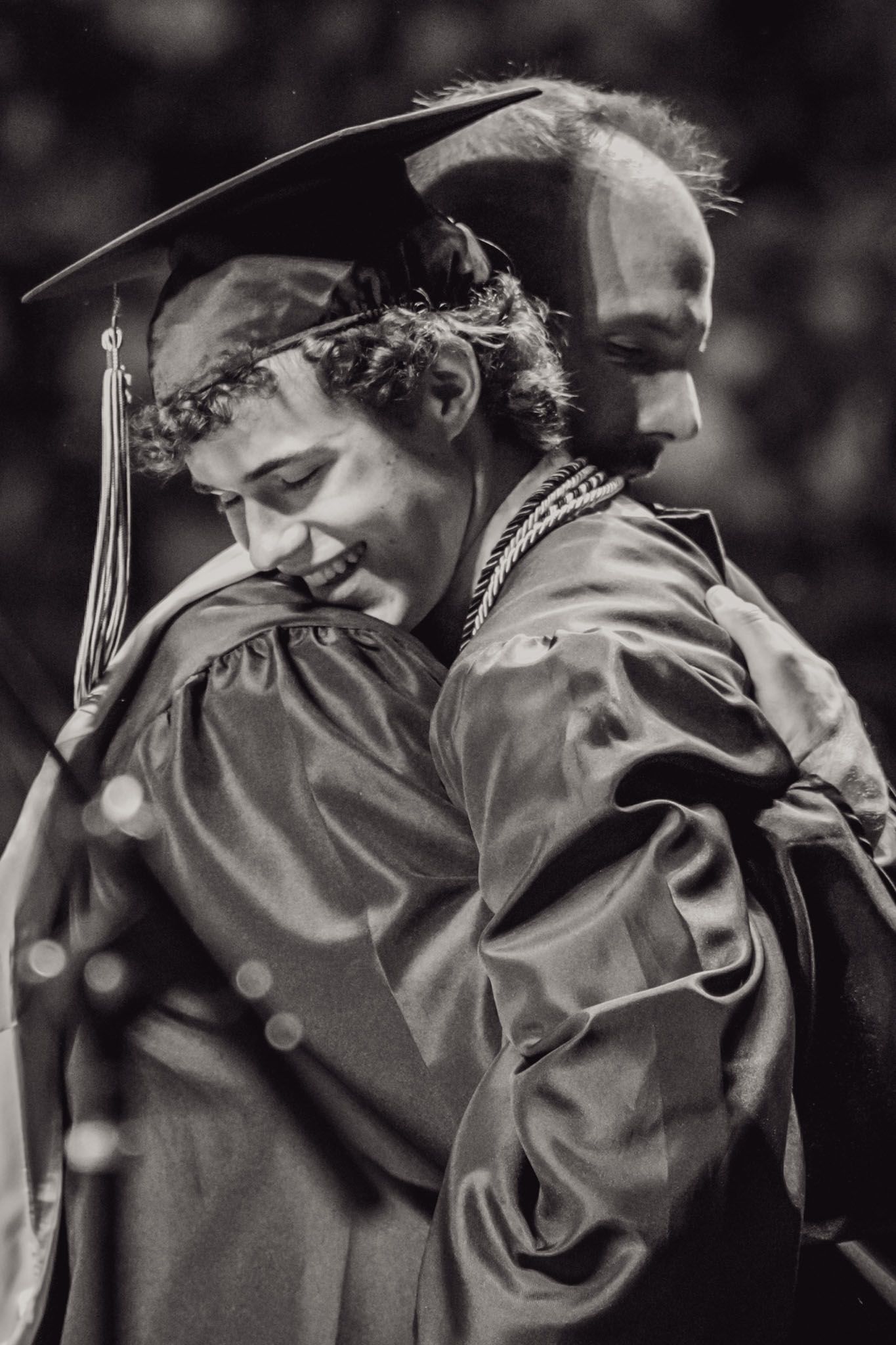 Students in graduation gowns hug and celebrate at commencement ceremony.