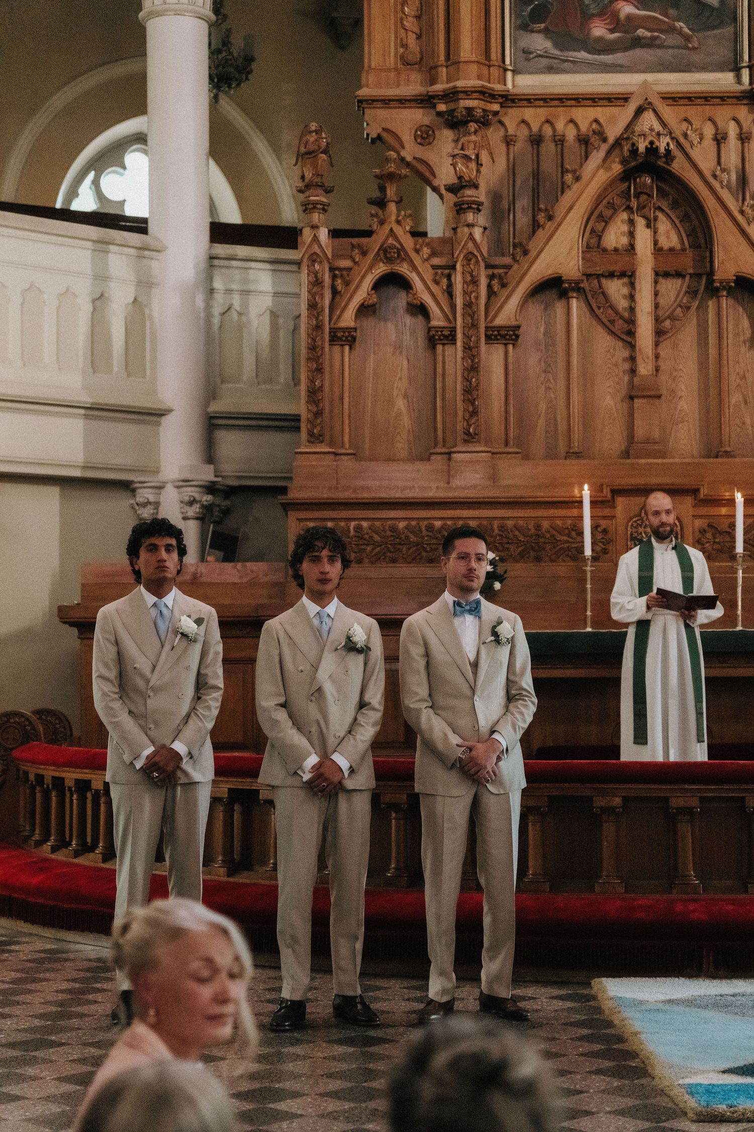 Three groomsmen in beige suits standing at the altar with a priest during a church wedding ceremony.