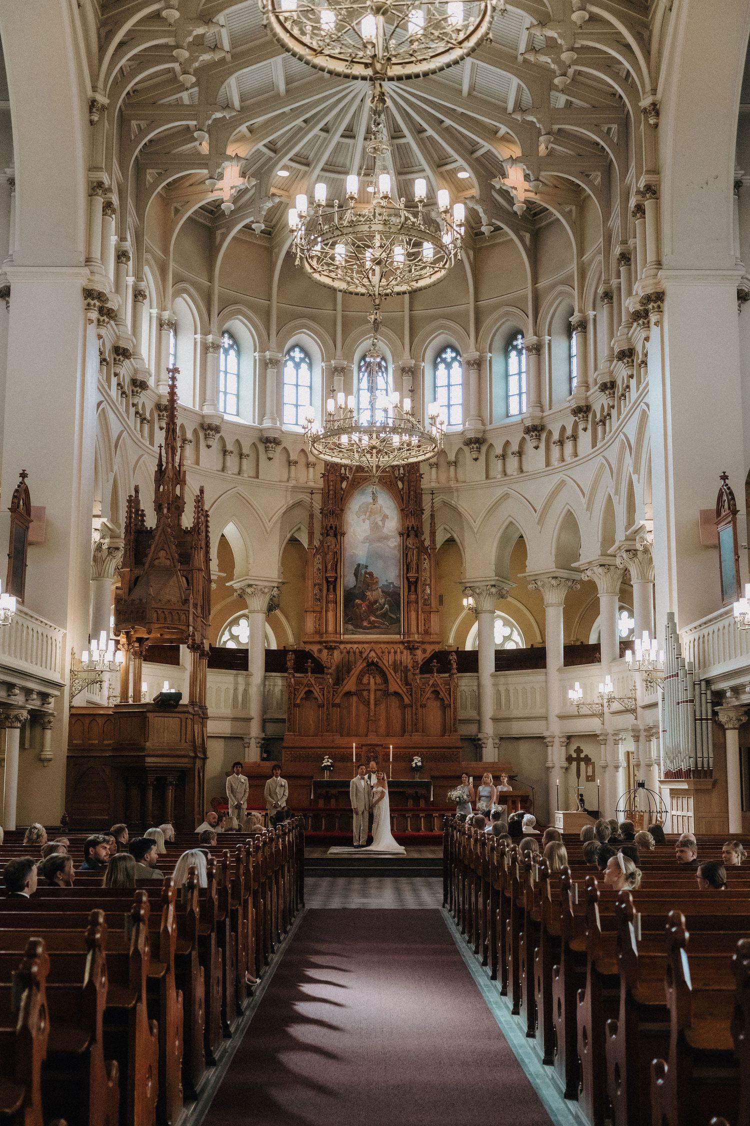 Elegant church wedding ceremony with ornate altar, chandeliers, and guests seated in wooden pews.