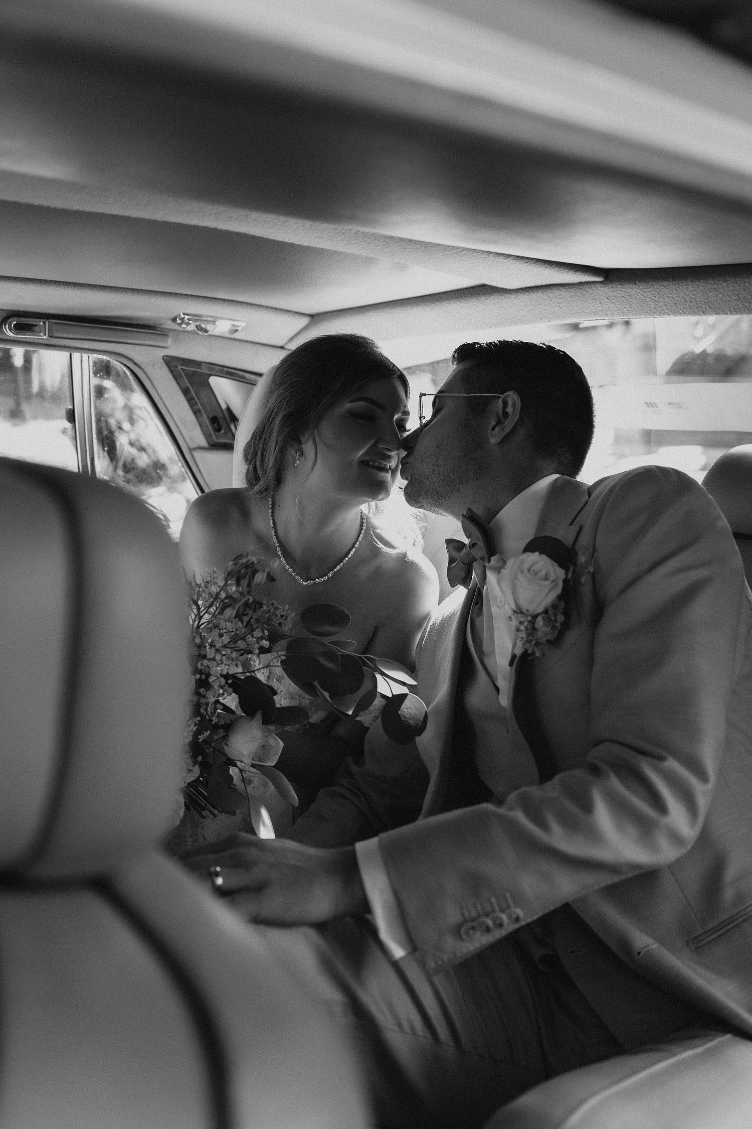 Black and white intimate photo of bride and groom sharing a kiss inside a vintage wedding car.