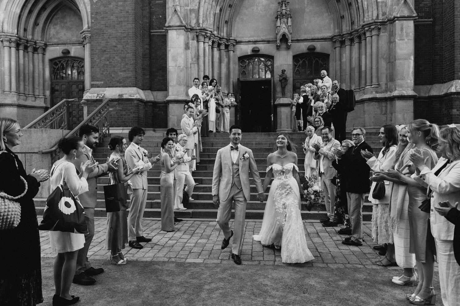 Black and white photo of bride and groom walking through cheering guests outside a Gothic church.