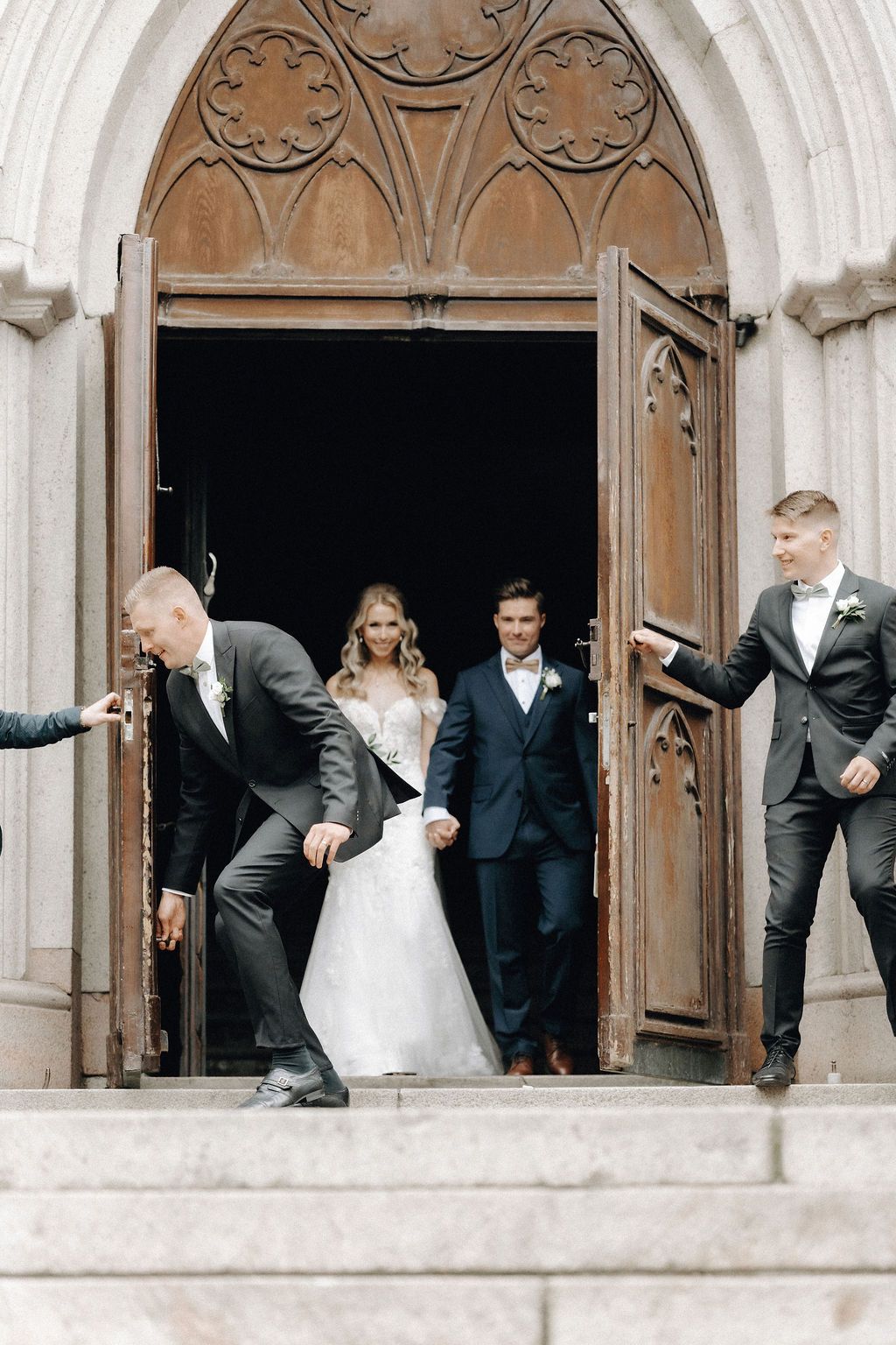 Bride and groom exit through ornate wooden Gothic church doors assisted by groomsmen in bow ties.
