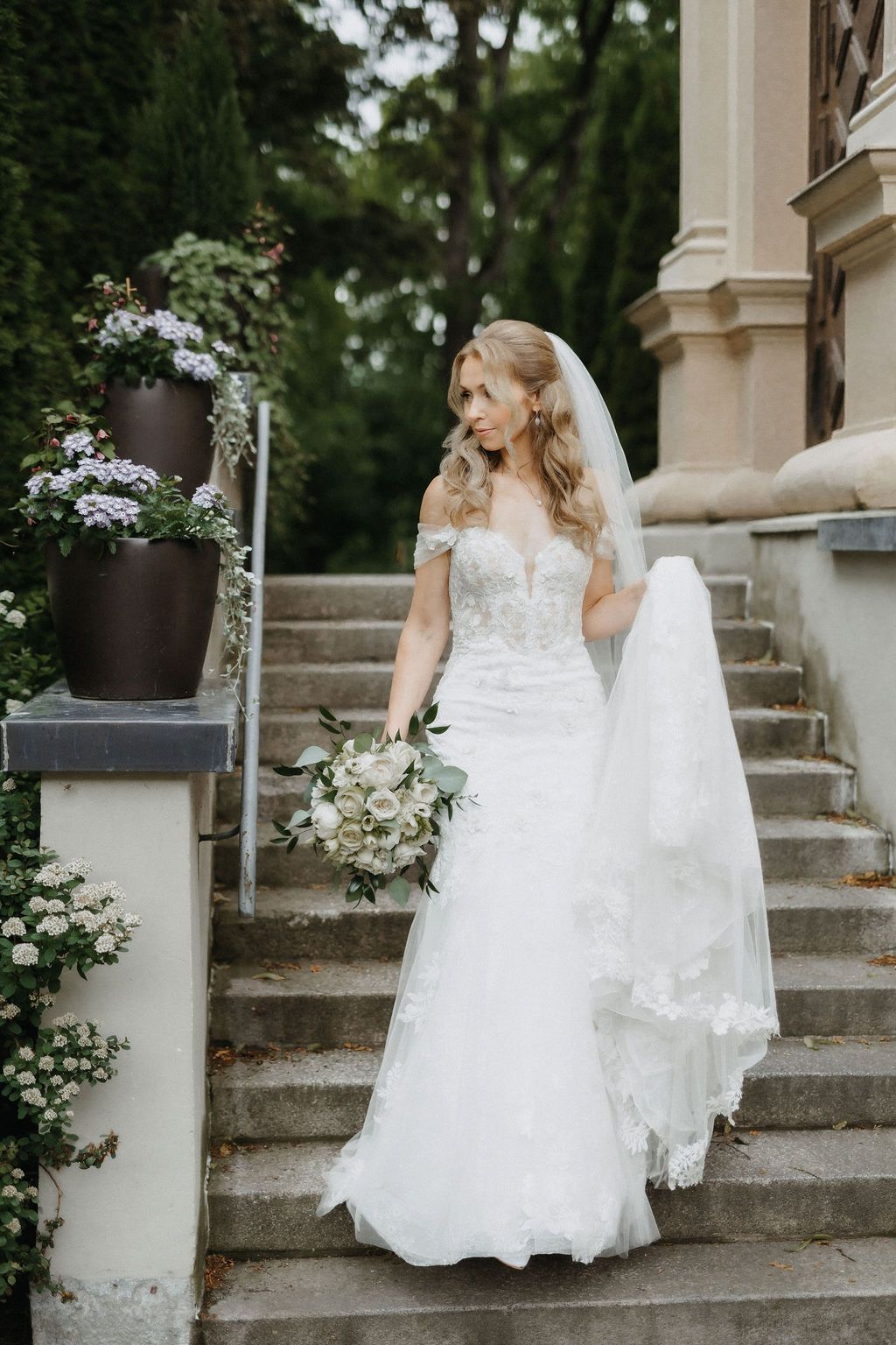 Blonde bride in off-shoulder lace wedding gown with flowing veil walks down stone steps of elegant venue.