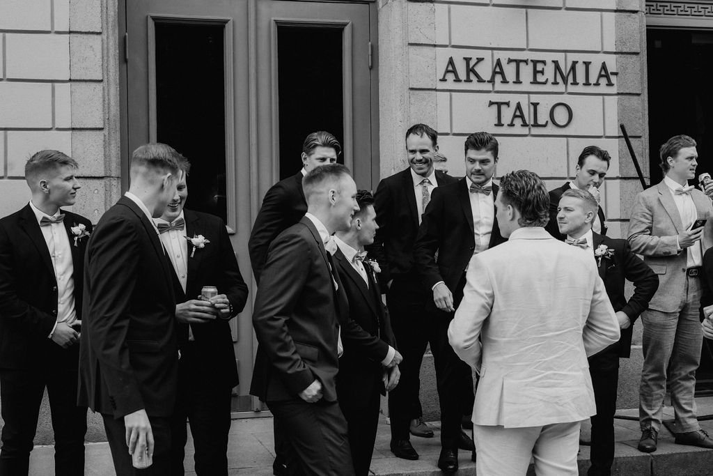 Groomsmen in black suits gather outside Akatemia Talo building in black and white wedding photo.