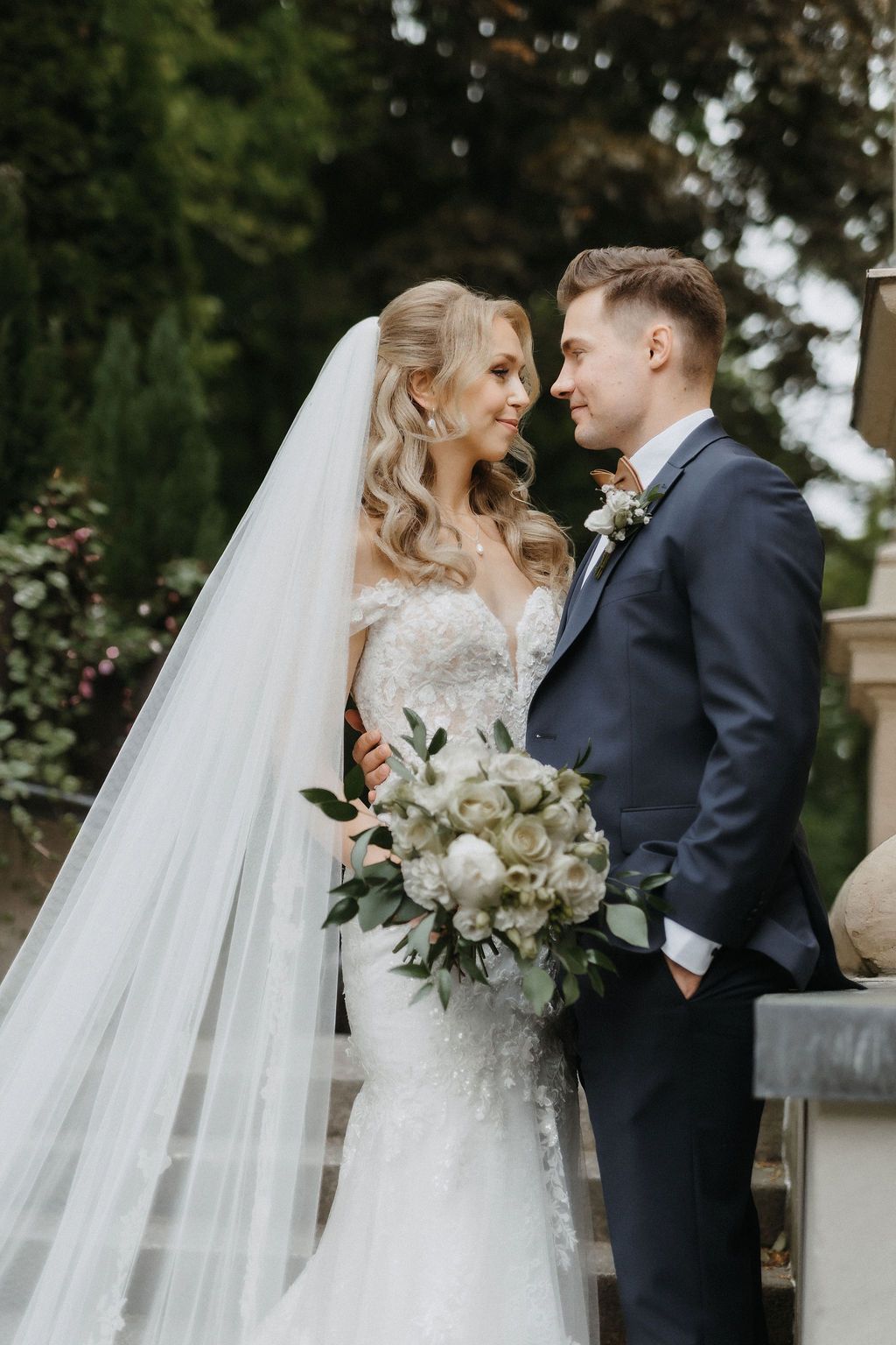 Bride and groom gaze at each other outdoors, bride in off-shoulder lace gown holding white peony bouquet.